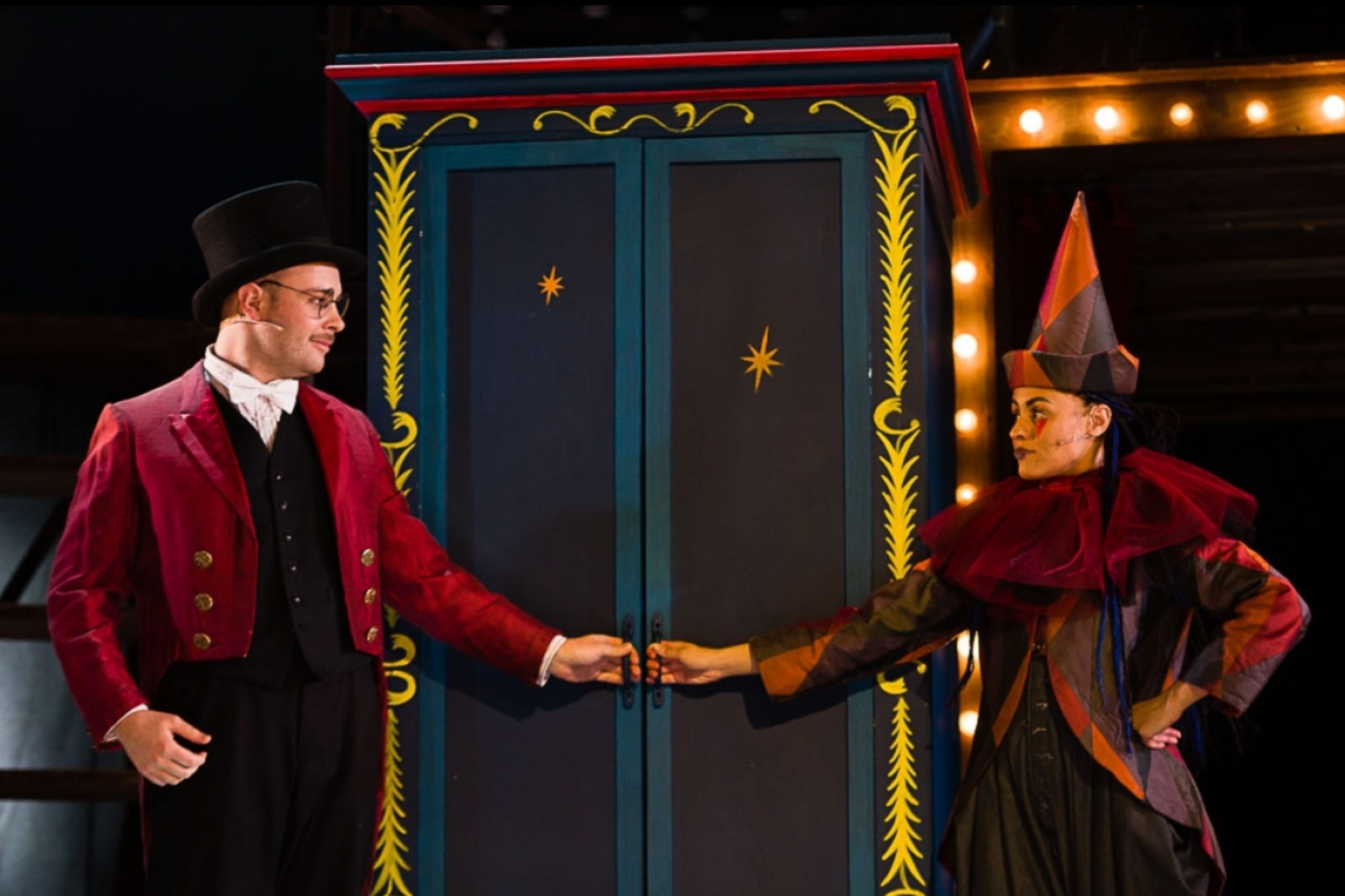 A man and a woman in theatrical costumes on stage, holding hands in front of a black door with stars, decorated with bright yellow and red trim, and surrounded by string lights.