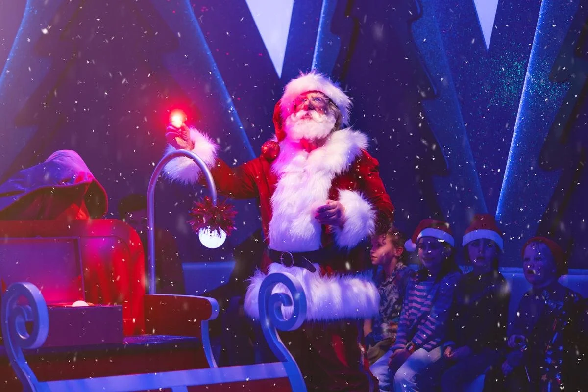 Santa Claus holding a glowing red object with children sitting nearby in Christmas hats, surrounded by falling snow and festive lights.