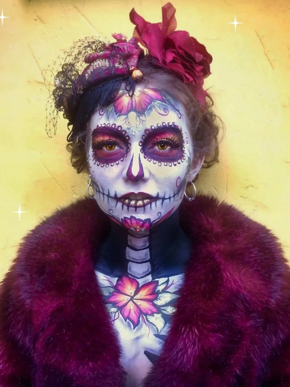 Woman dressed in Día de los Muertos style makeup with colorful floral and skeleton designs, wearing a dark fur coat and large red flower headpiece, standing against a yellow wall.