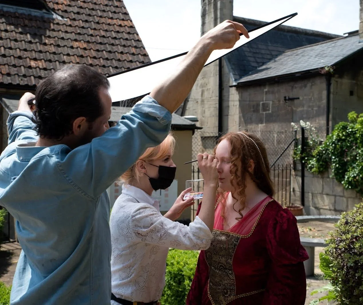A woman with curly red hair is getting her makeup done outdoors by two makeup artists, one holding a palette, while another holds a sheet of paper over her head to shield her from the sun.