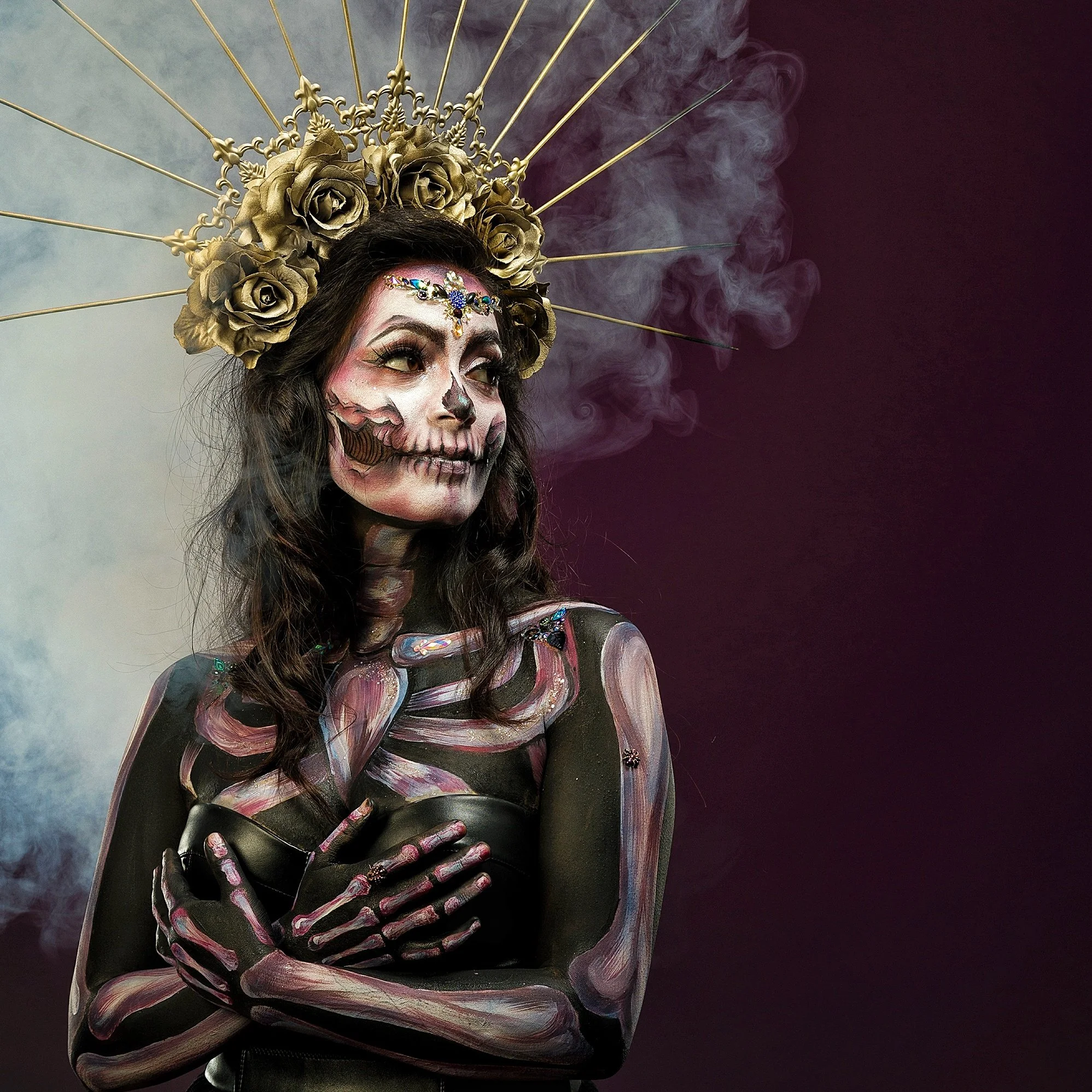 Woman with skeleton face paint and body art, wearing a headdress with gold roses and radiating gold spikes, standing with arms crossed and surrounded by purple and smoky background.