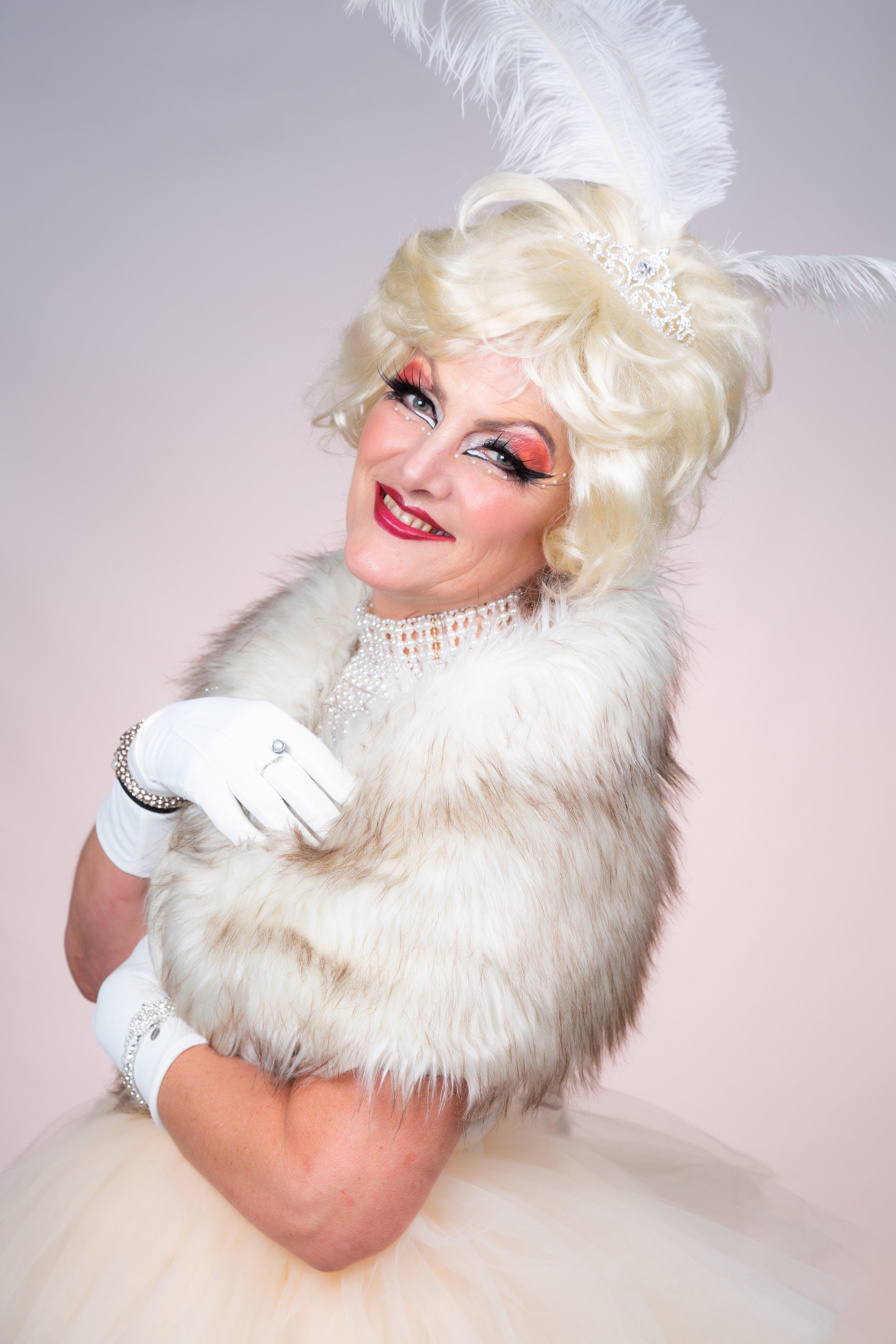 Portrait of a smiling woman in an elegant white gown, fluffy fur shawl, pearl choker, white gloves, and a large feathered headpiece, set against a neutral background. Drag makeup. Pastel colours. Painted Peach