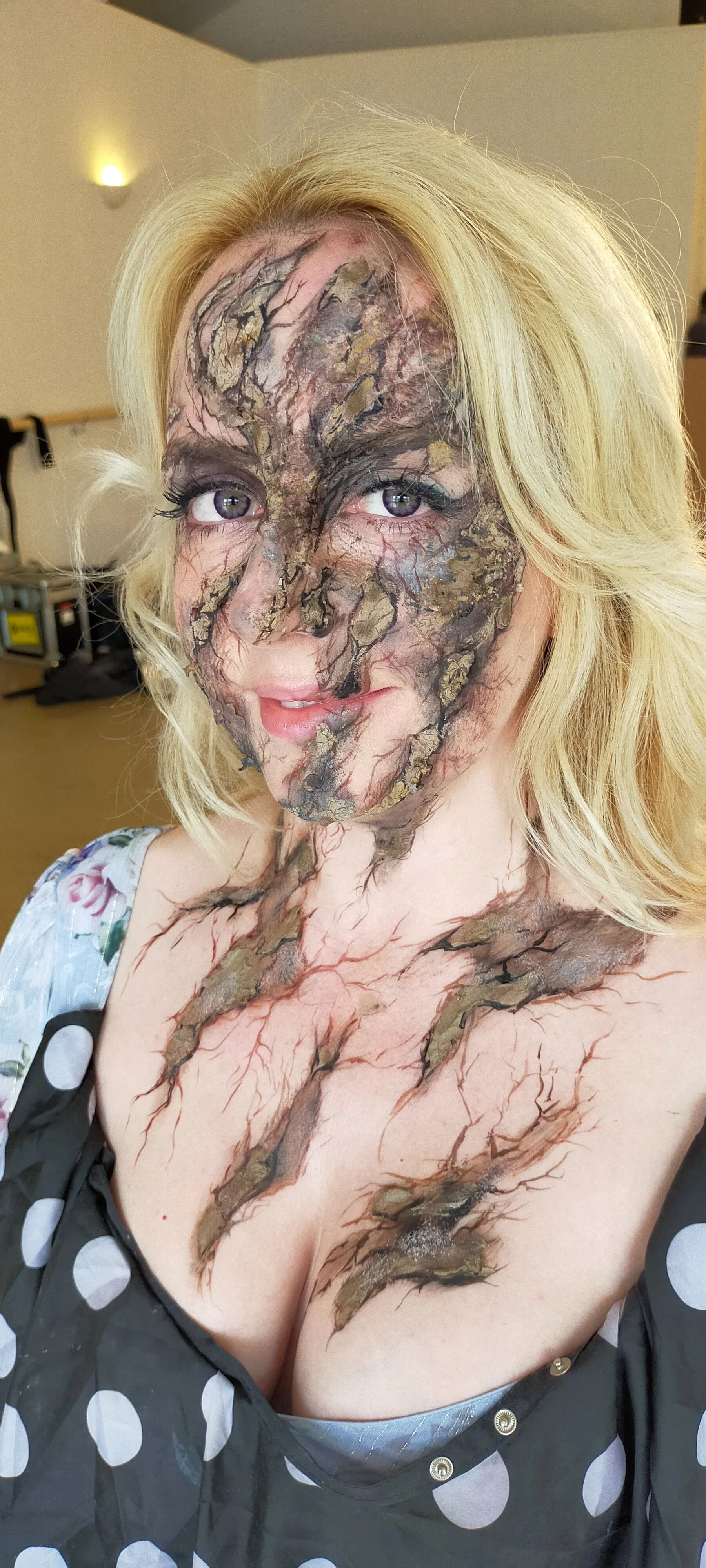 Woman with artistic makeup resembling cracked earth or bark on her face, neck, and chest, wearing a polka dot dress, in a room with plain walls and equipment in the background.