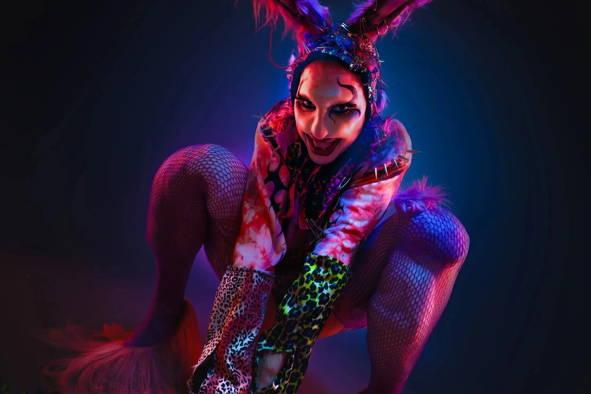 A woman dressed in colorful, punk-inspired costume with fishnet stockings, leopard print gloves, and spikes, crouching with a playful expression, illuminated by vibrant blue and pink lighting.