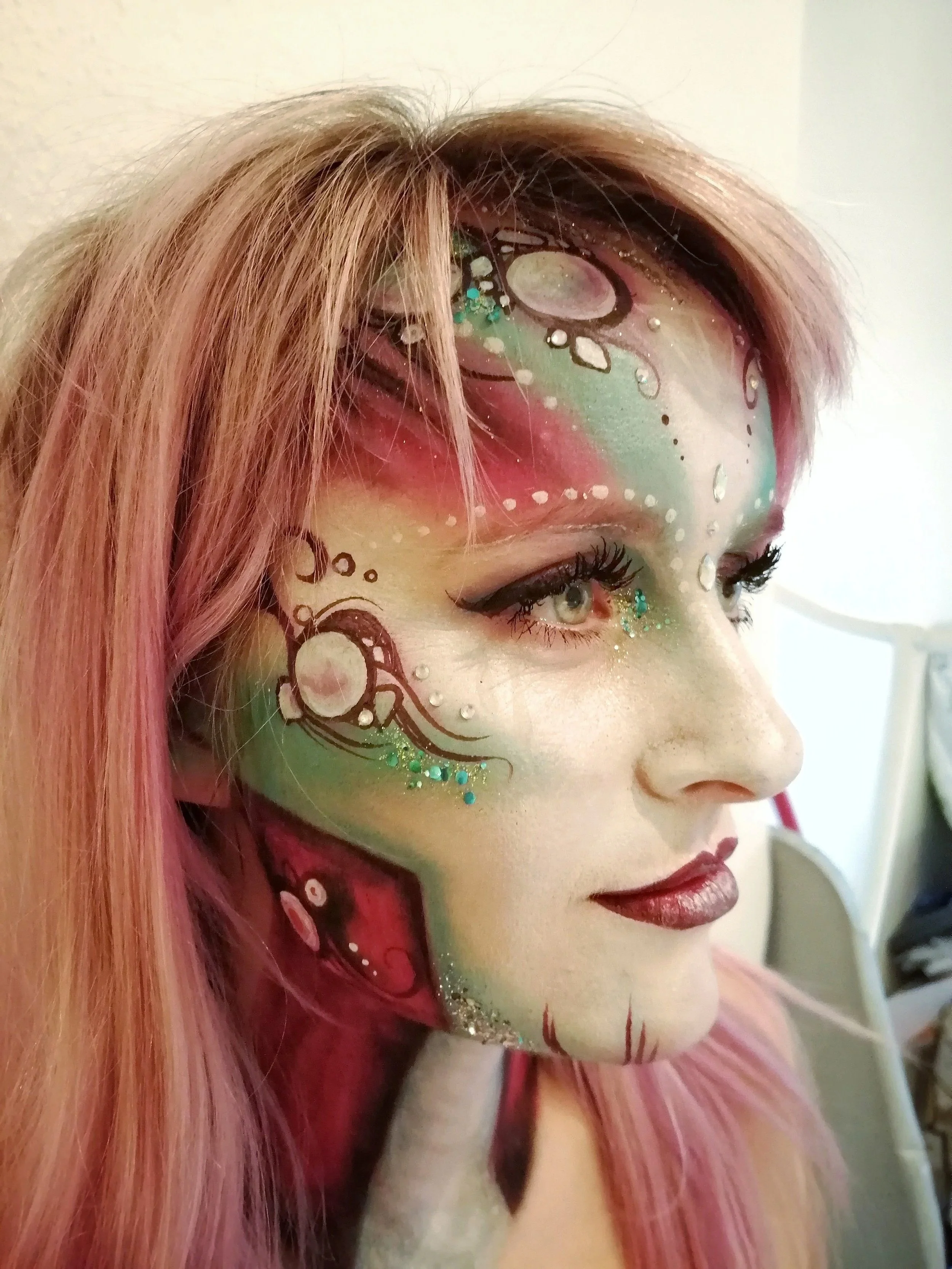 A close-up of a woman's face with intricate, colorful fantasy makeup featuring bubbles, swirls, and glitter.
