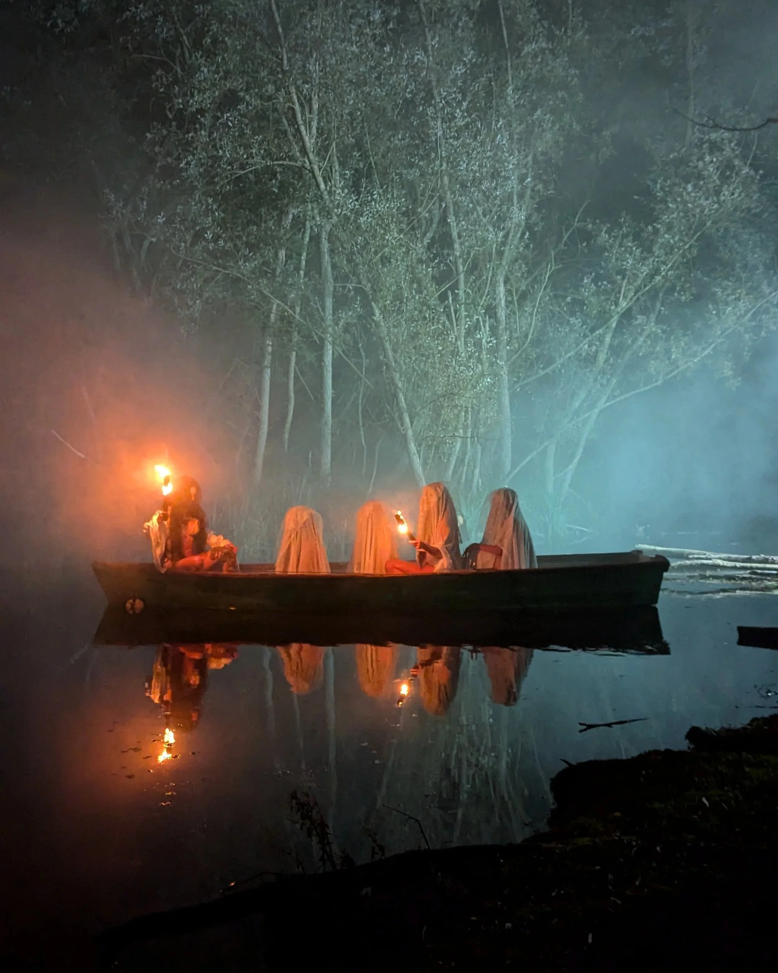 A boat carrying four people with white cloth coverings on their heads, holding torches, floating on a calm body of water at night, with a wooded background and mist or fog.