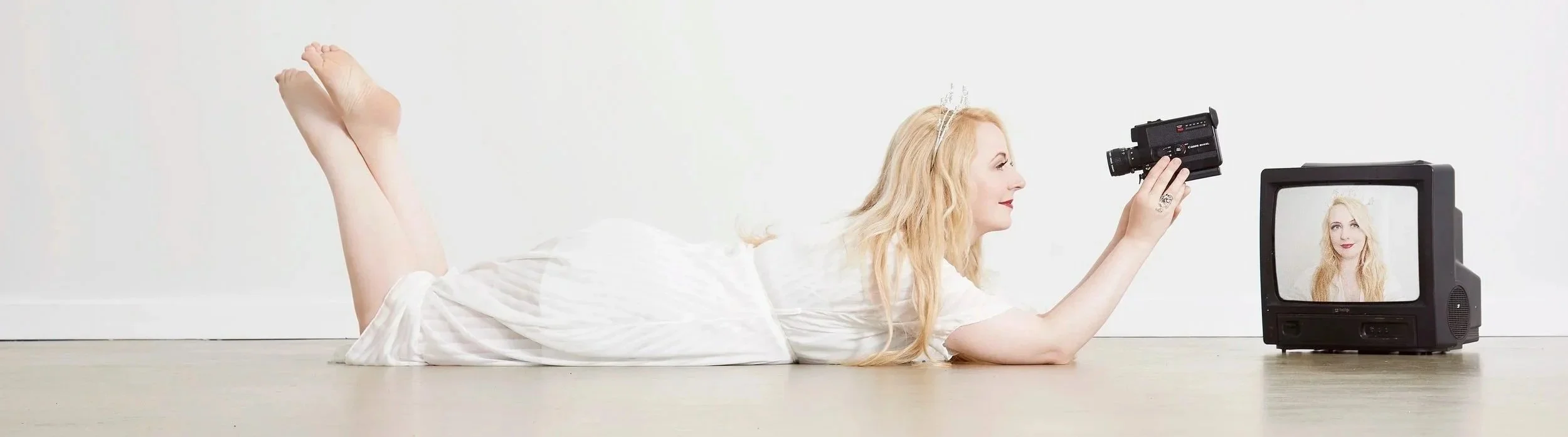 A woman lying on her stomach on the floor in front of a vintage television, filming herself with a handheld camera, with her reflection visible in the TV screen.