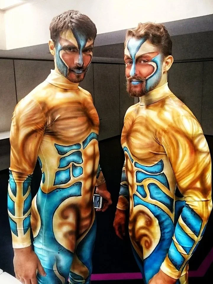 Two men with elaborate body and face paint resembling fantasy characters, standing indoors with dark walls in the background.