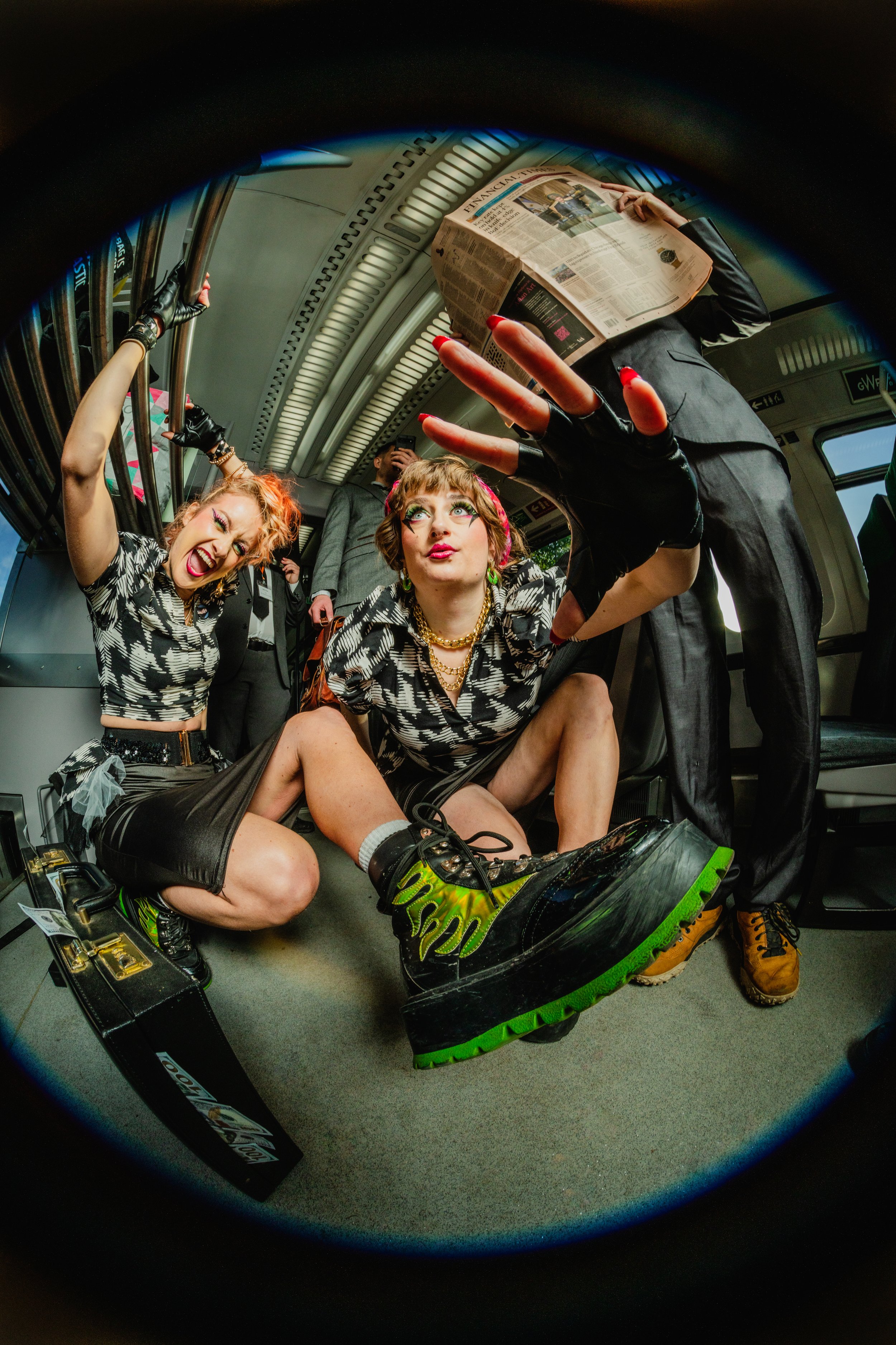 Two women and one man dressed in fashionable, edgy clothing, riding the floor of a subway train. The women are crouched down with one holding onto a metal handrail and the other taking a selfie, while the man is standing and reading a newspaper.