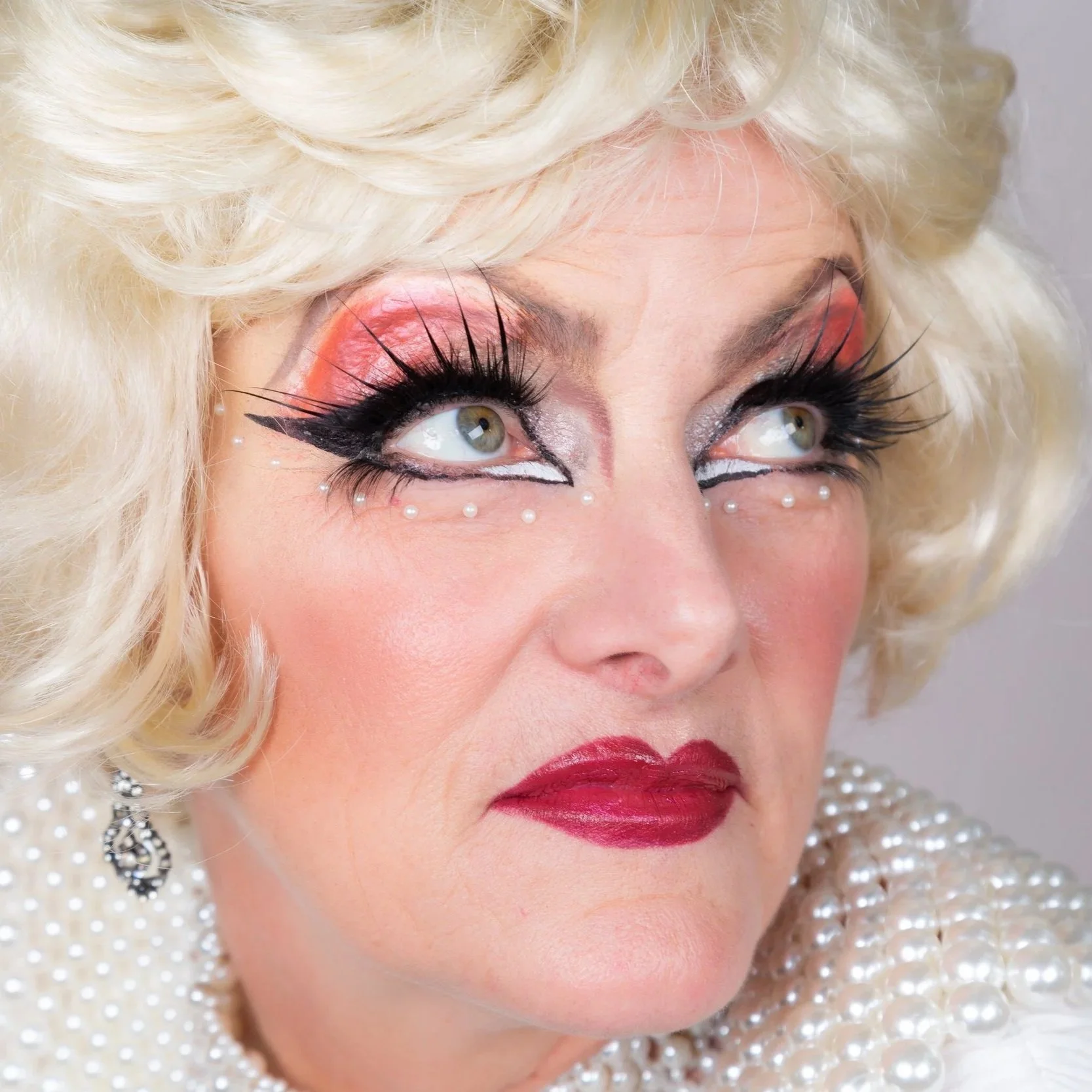Close-up of a woman with dramatic, colorful makeup resembling a clown or theatrical style, wearing a pearl necklace and earrings, with platinum blonde hair.