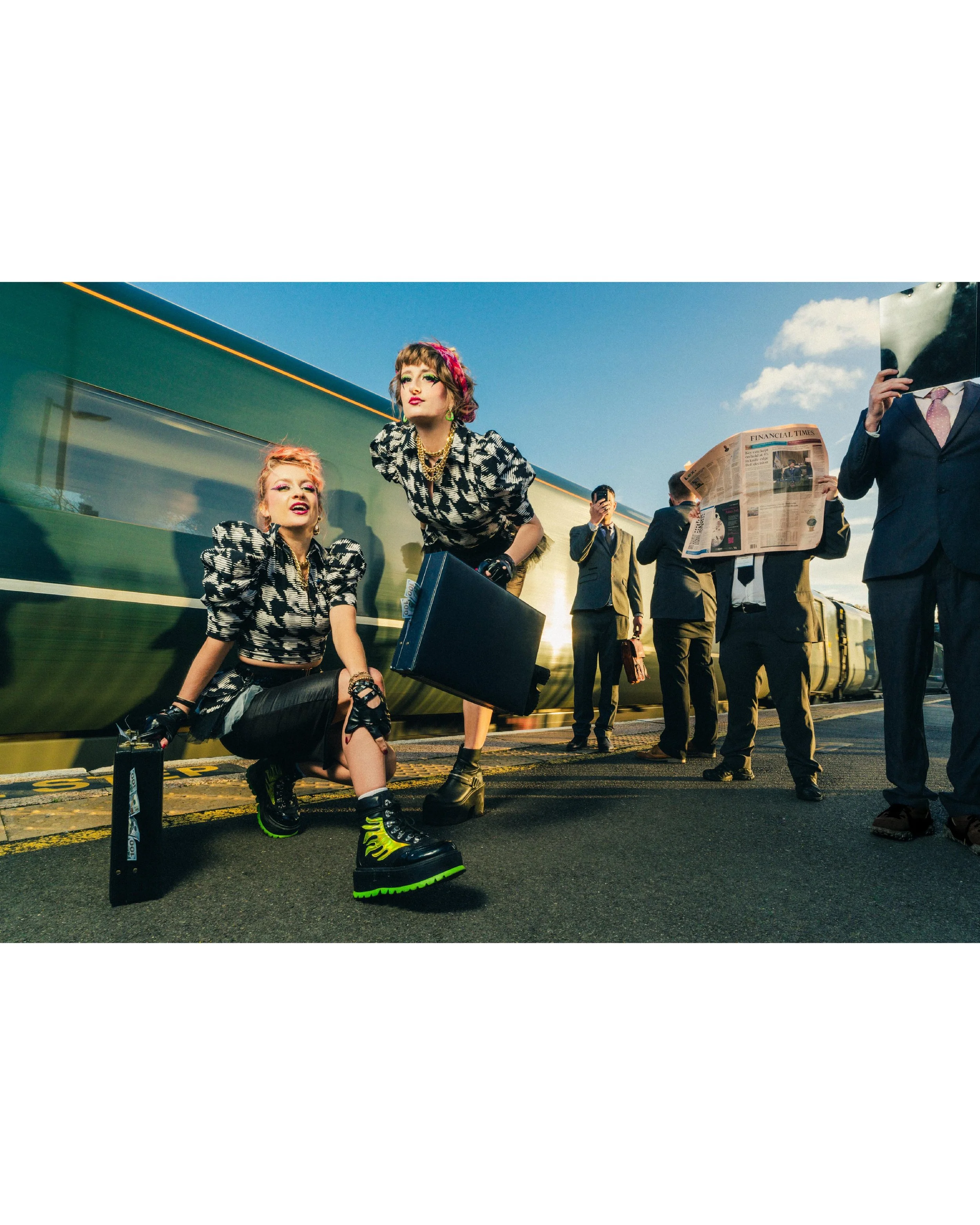 Fashionably dressed women at a train station with a moving train and men in suits reading newspapers in the background.