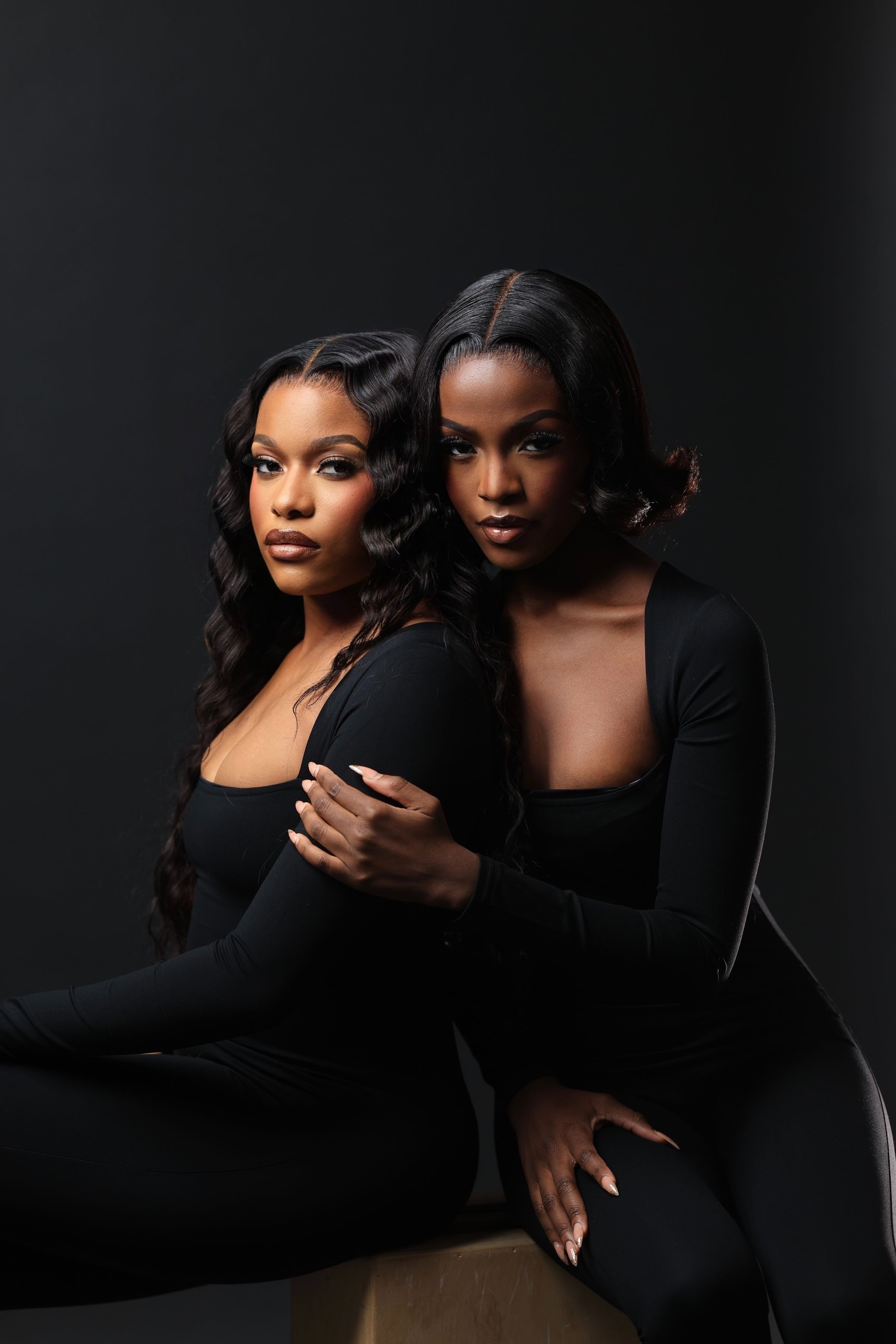 Two women with dark hair and makeup, wearing black long-sleeve outfits, posing closely together against a dark background.