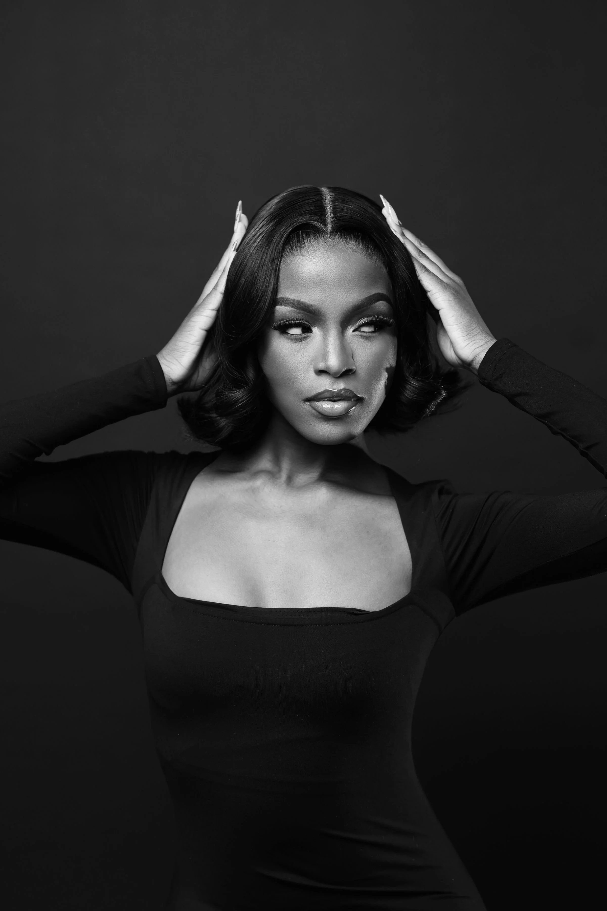 Black and white portrait of a woman with styled hair, wearing a long-sleeved top, with hands on her head, against a dark background.
