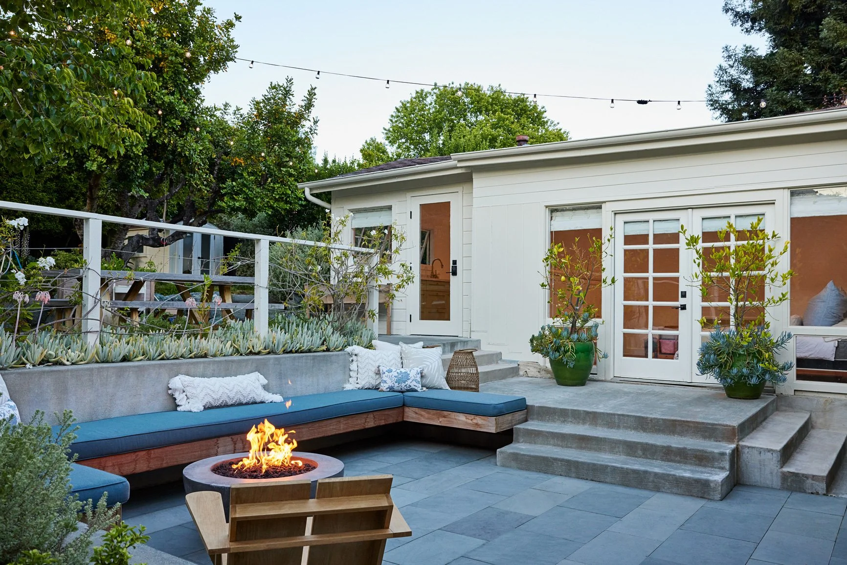 Example of an addition. Backyard patio with built-in blue cushioned bench around a fire pit, potted plants, and a white ADU with large glass-paneled doors, surrounded by trees and string lights