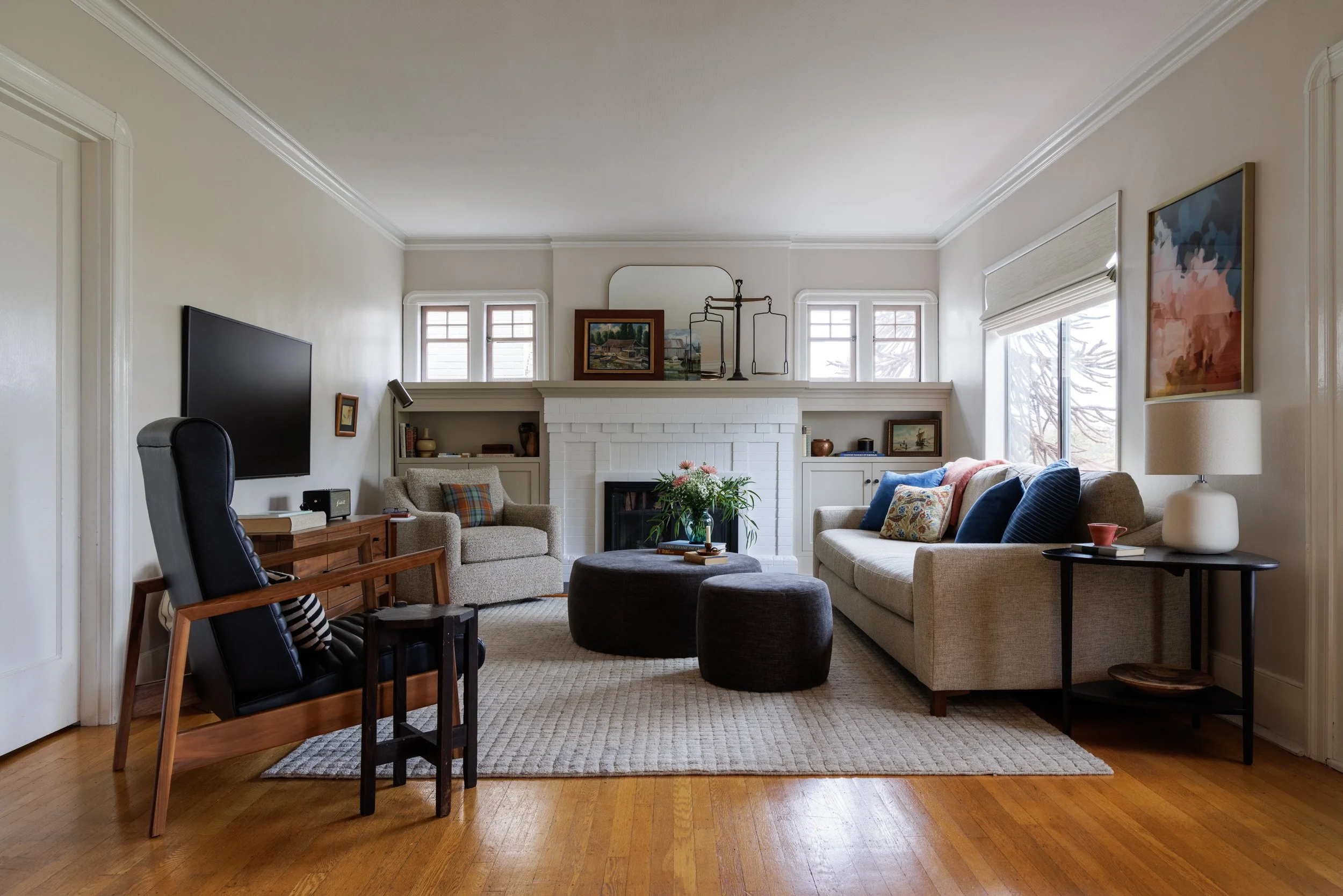 Living room with beige sofa, armchairs, dark gray ottomans with flowers and books on top of one, TV over console table, fireplace, windows with blinds, framed artworks, side table with lamp and decorative bowl, hardwood floor with area rug.
