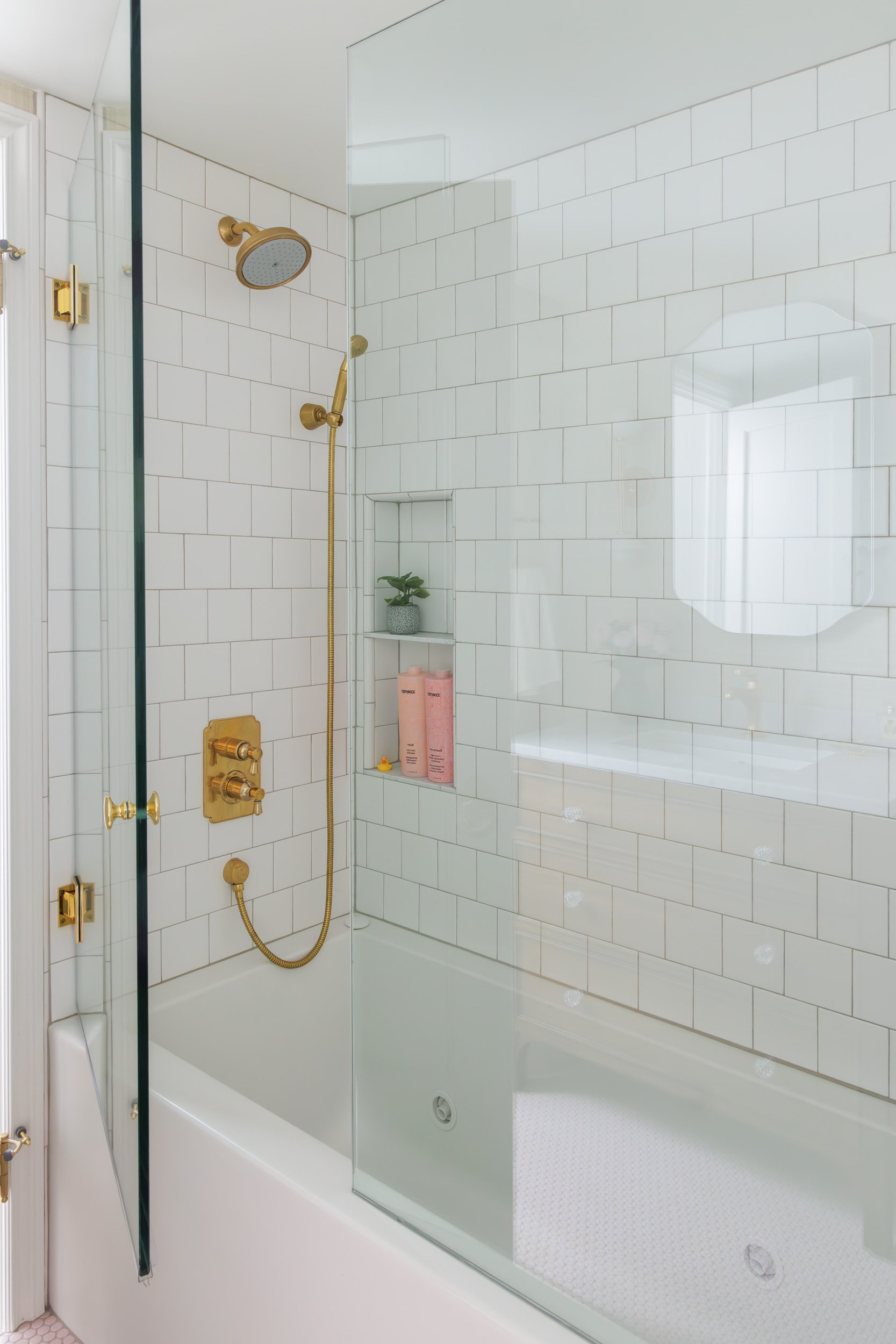 Modern bathroom shower with white subway tiles, gold fixtures, and a glass door. Includes a built-in shelf with a small plant and pink bottles, and a shower head with a handheld attachment.