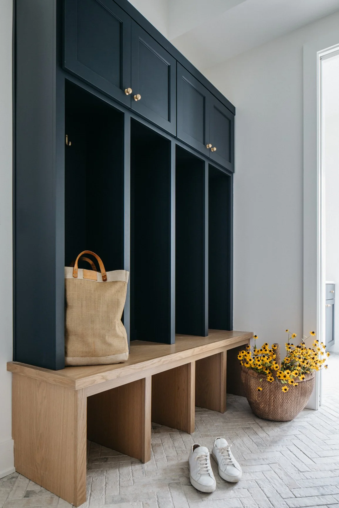A modern entryway with a dark blue built-in storage unit featuring upper cabinets and open cubbies. A beige tote bag rests on a wooden bench below, alongside a large woven basket filled with yellow flowers and a pair of white sneakers on the floor. Natural light streams in from a nearby door.