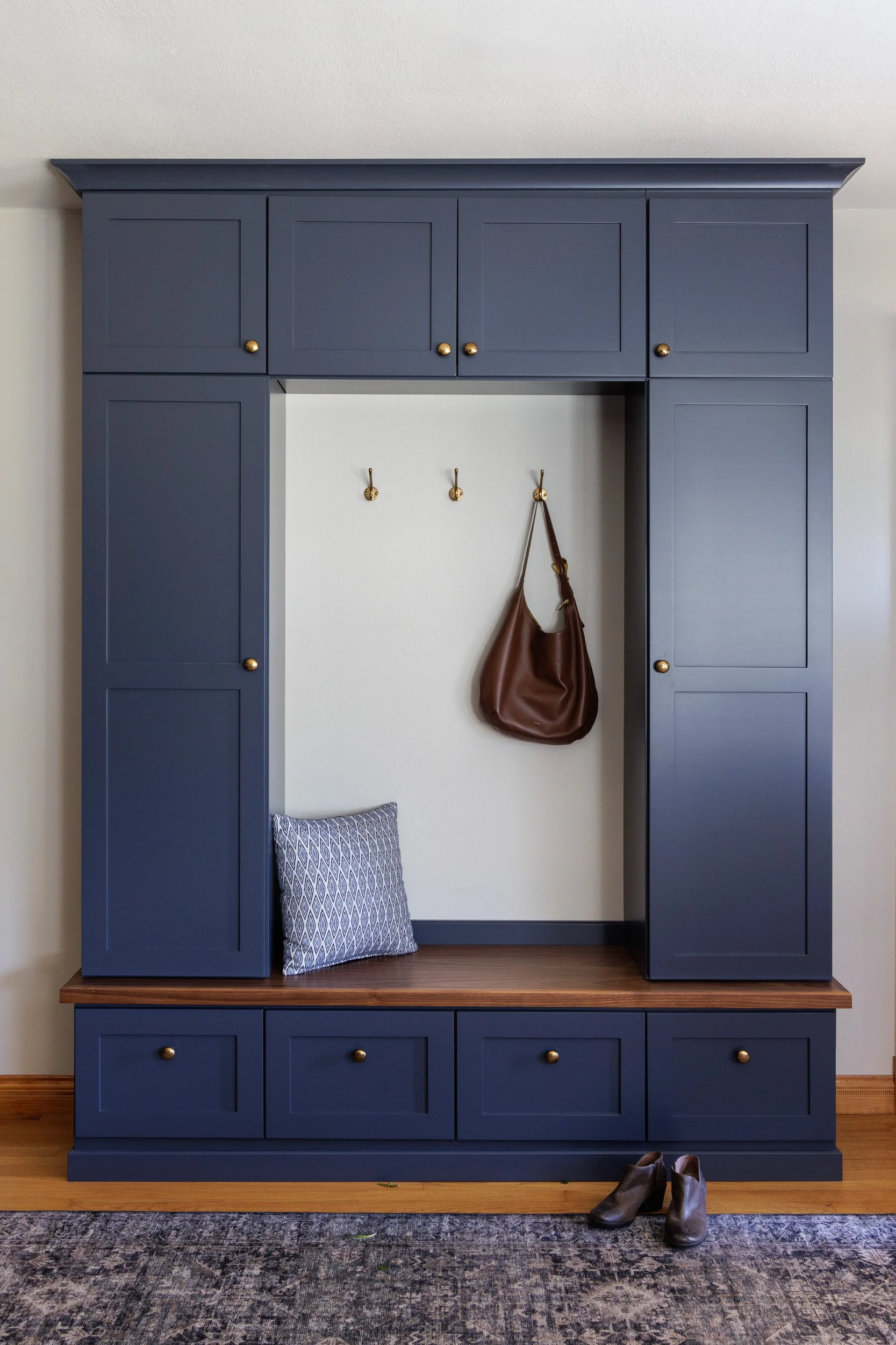 A navy blue entryway with built-in storage and three hooks on a white wall, a brown handbag hanging, a blue patterned pillow on the bench, and a pair of brown shoes on a rug on the floor.