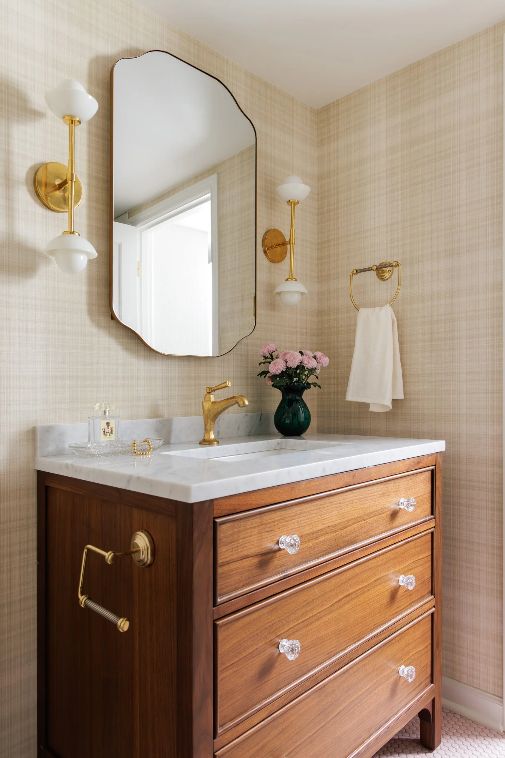 Vintage bathroom vanity with a marble top, brass fixtures, a mirror, wall sconces, and pink flowers in a green vase.