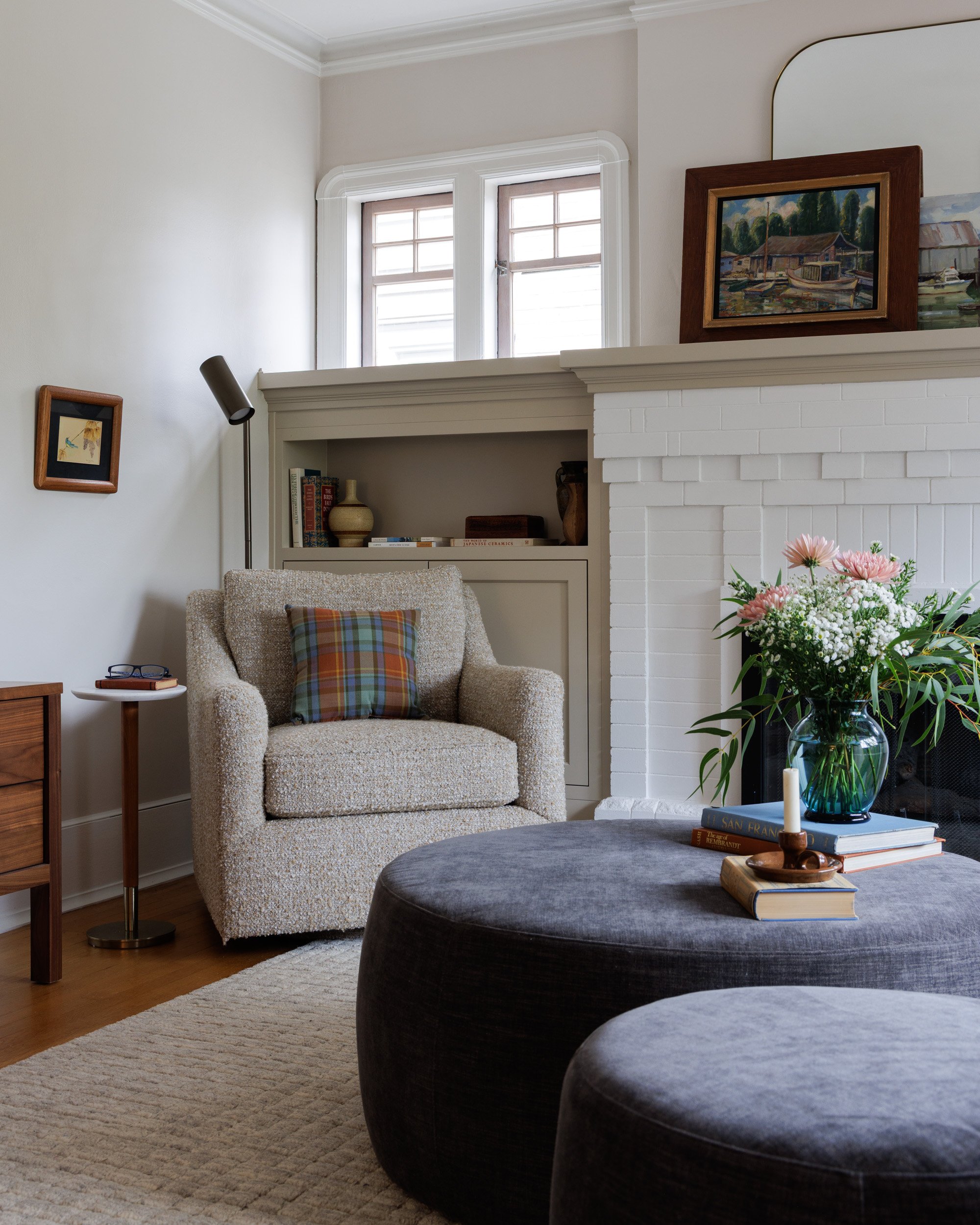 A living room with a white fireplace, armchair with a plaid pillow, side table with glasses, wall art, reading lamp, dark gray ottomans with flowers and books on one, a white brick fireplace with mirror and art above, and a window with white trim.