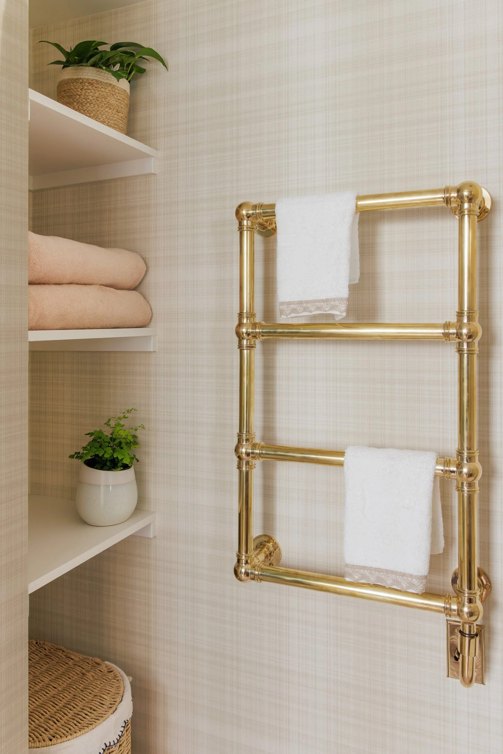 A bathroom with beige plaid wallpaper, a white shelf with light pink towels and a potted plant, and a gold towel warmer holding two white towels.