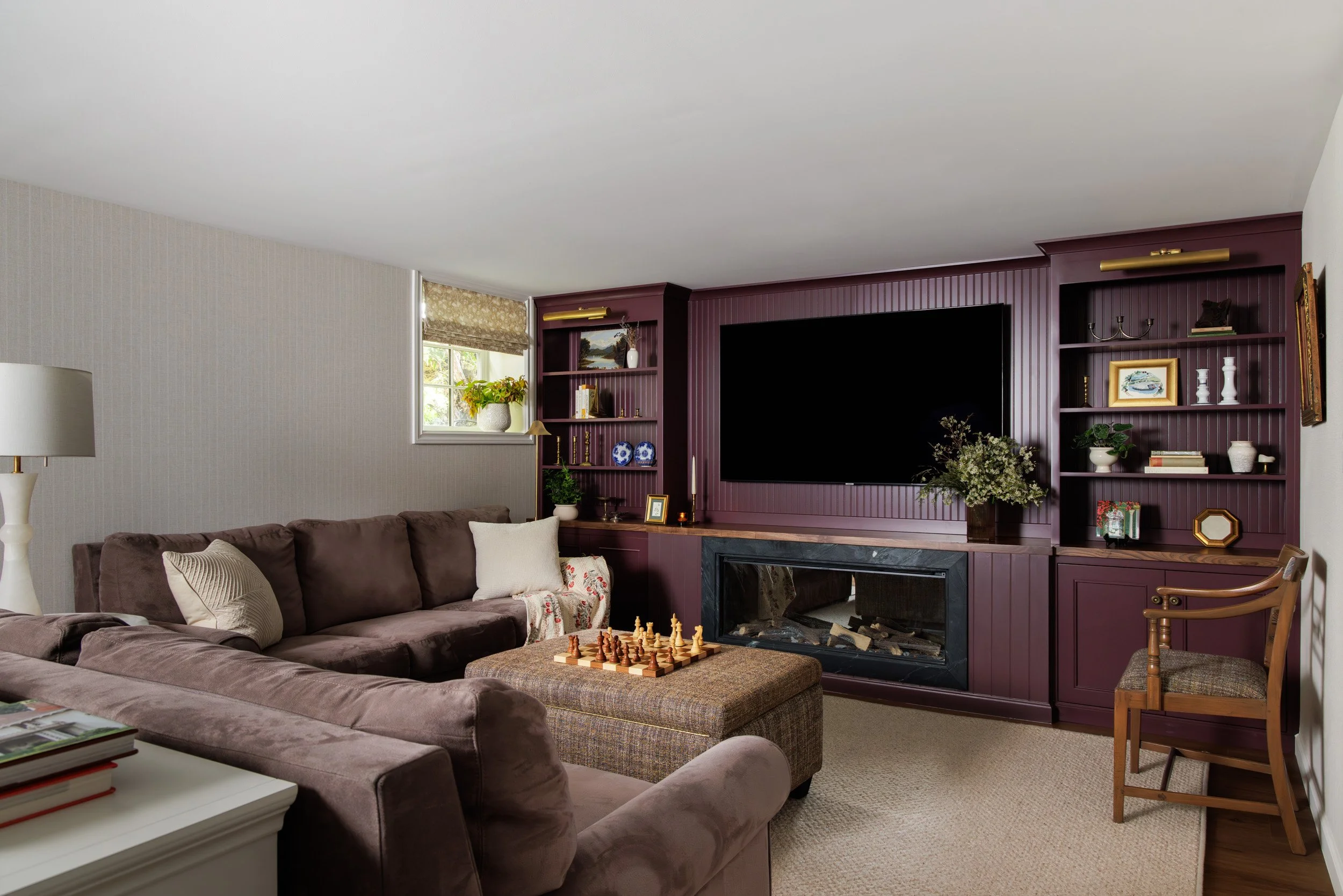 Living room with brown sectional sofa, lamp to the left, fireplace, purple built-in shelves filled with decor, large flat-screen TV, window with floral roman shade, decorative plants, an ottoman with a chess set on it, and a wooden chair.