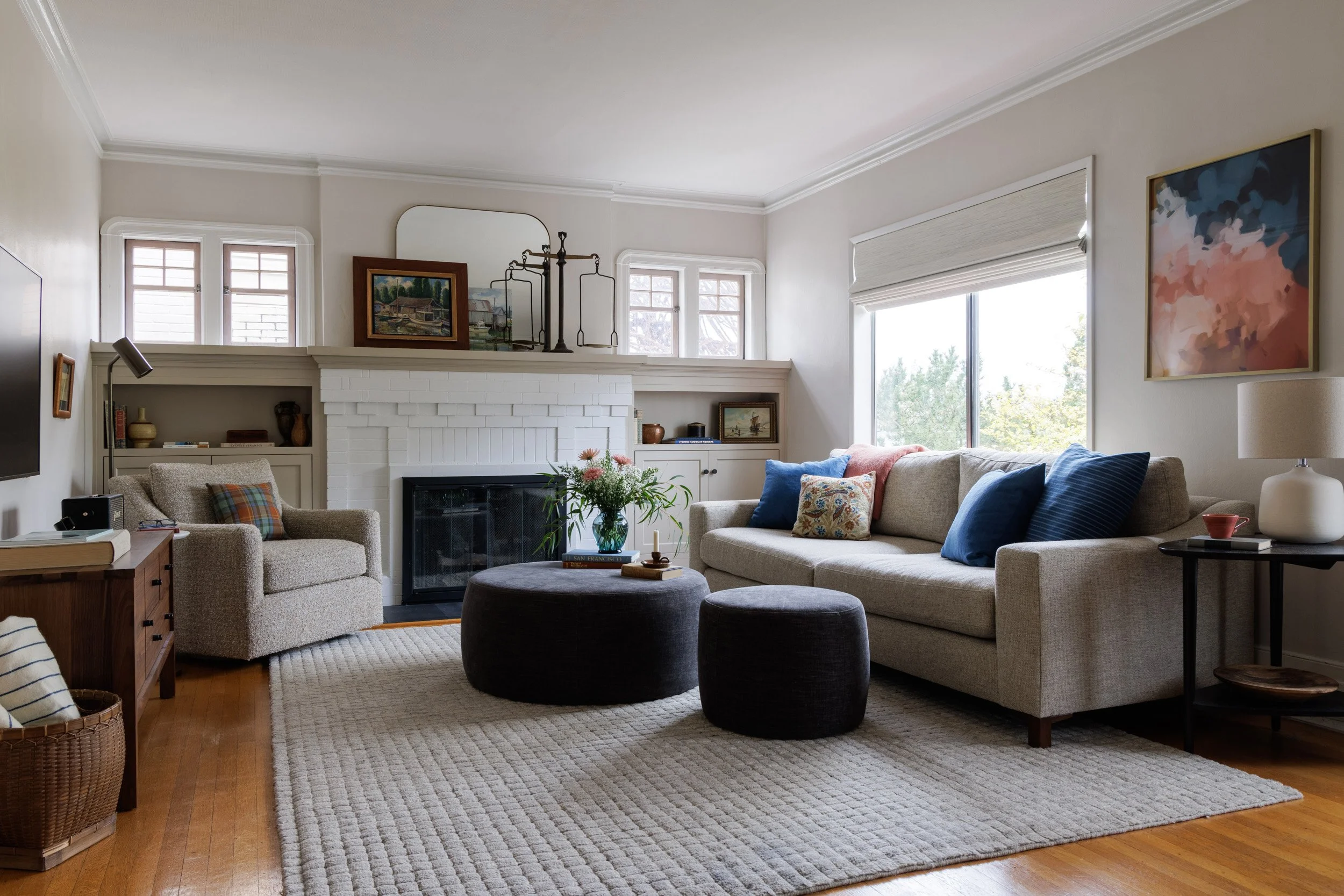 Living room with beige sofa, armchair, dark gray ottomans with flowers and books on top, white brick fireplace with art and mirror above, built-ins with decor, large window, side table with lamp, mug and bowl, wall art, hardwood floor with area rug.