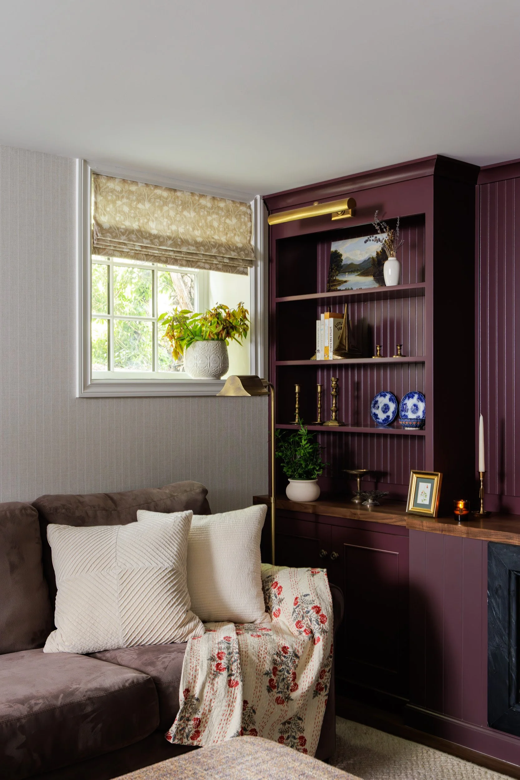 A living room corner with a window, a brown sofa with white pillows and a floral blanket, and a purple built-in bookshelf filled with decorative items, books, and plants.