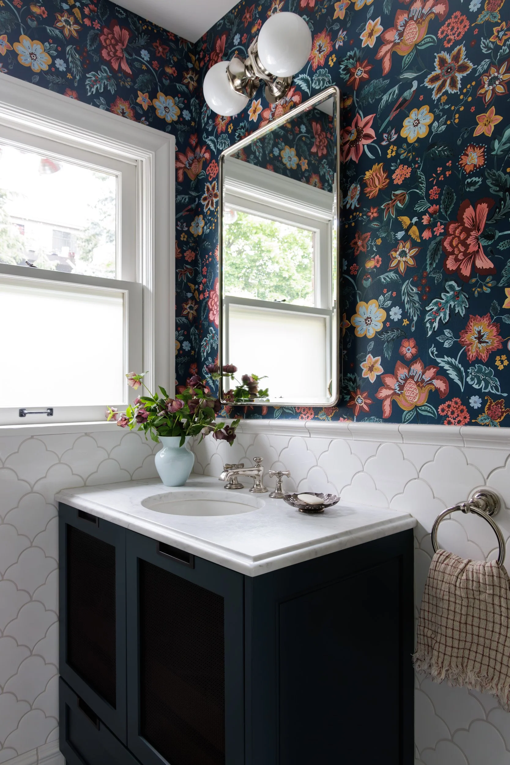 A bathroom with dark floral wallpaper, a white sink vanity, a large rectangular mirror, a vintage-style light fixture with round bulbs, a window with white trim, a vase with purple flowers, a small dish, and a beige towel hanging on a silver towel ri