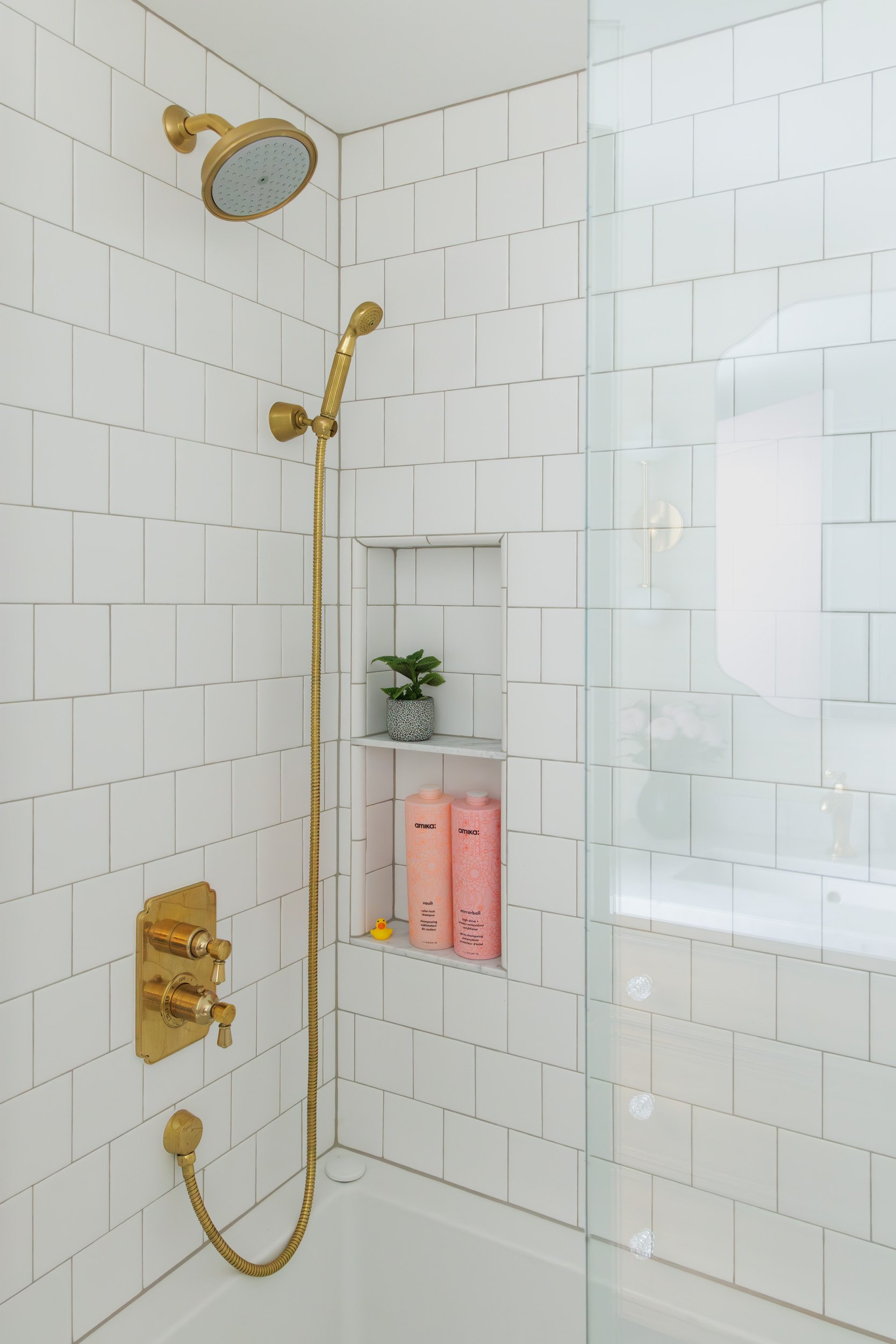 Modern bathroom shower with white subway tiles, gold fixtures and a shower head with a handheld attachment. Includes a built-in shelf with a small plant, pink bottles, and tiny rubber duck.