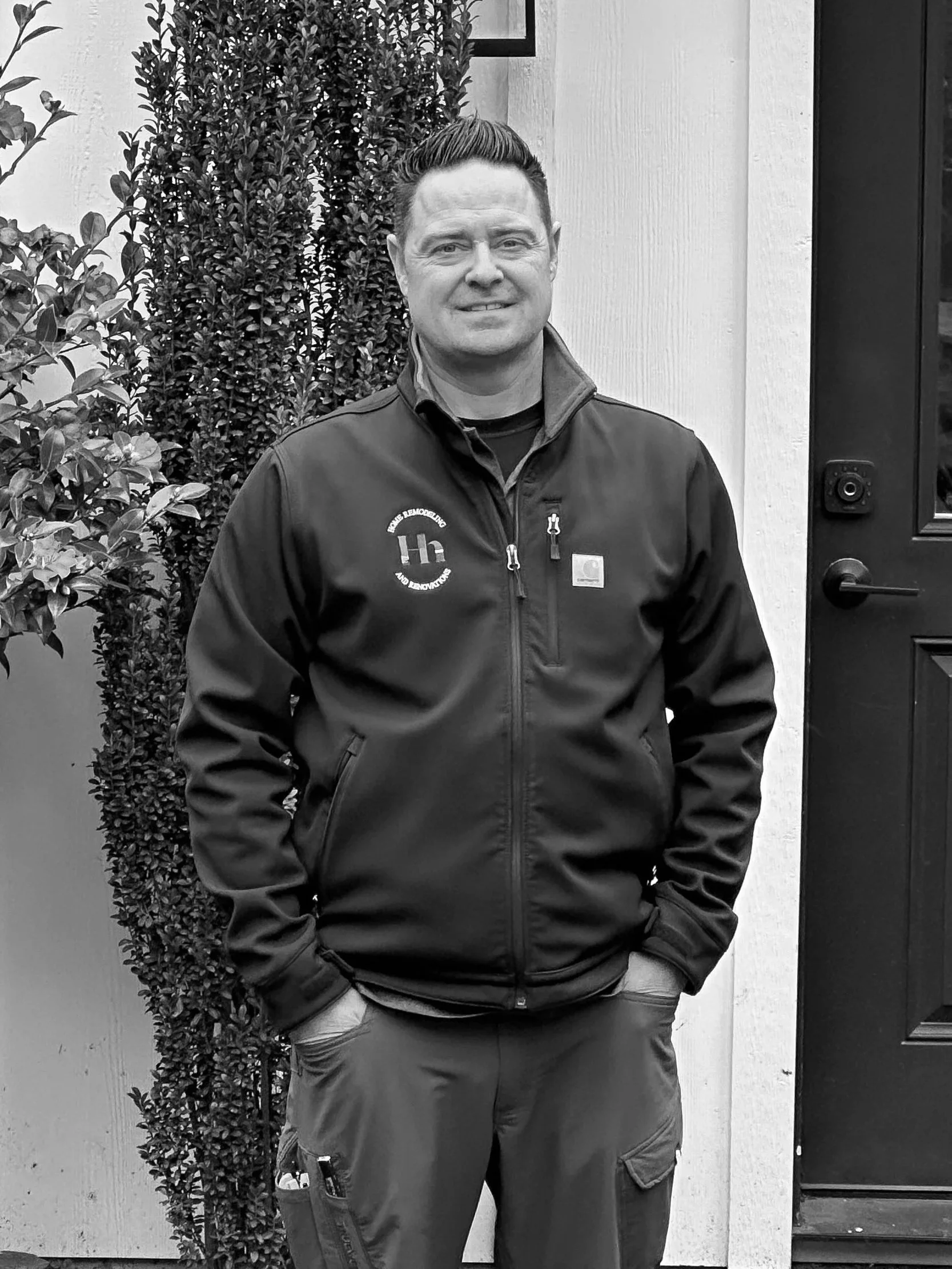 The about photo for Haberdash Home founder Troy Grindle. He is standing outdoors with hands in pockets, wearing a jacket with his logo, in front of a white wall, a door, and bushes.