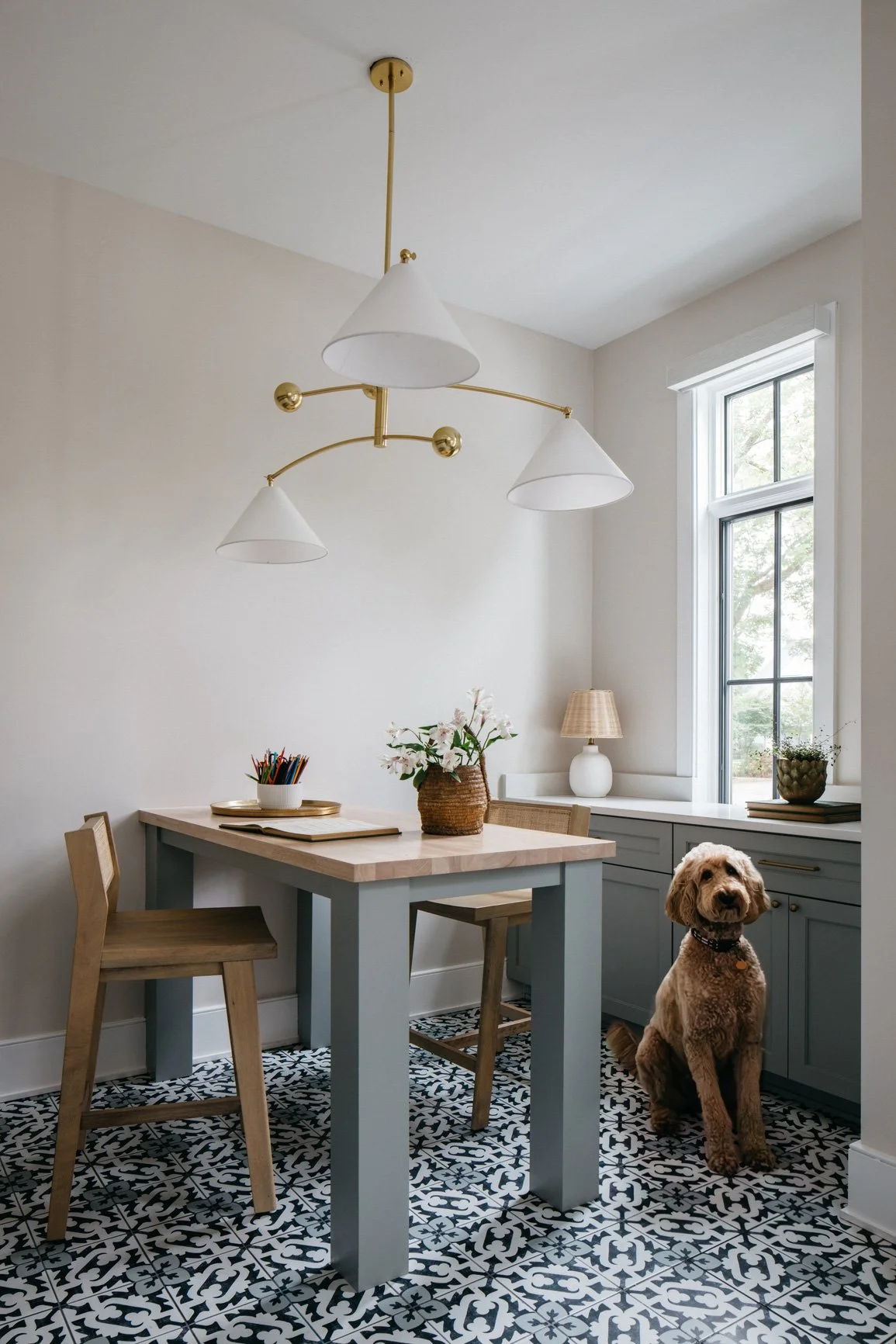 A homework station with a light-colored table, two wooden chairs, a dog sitting on patterned blue-and-white tile flooring, a window with natural light, and decor including a vase with flowers and a lamp.