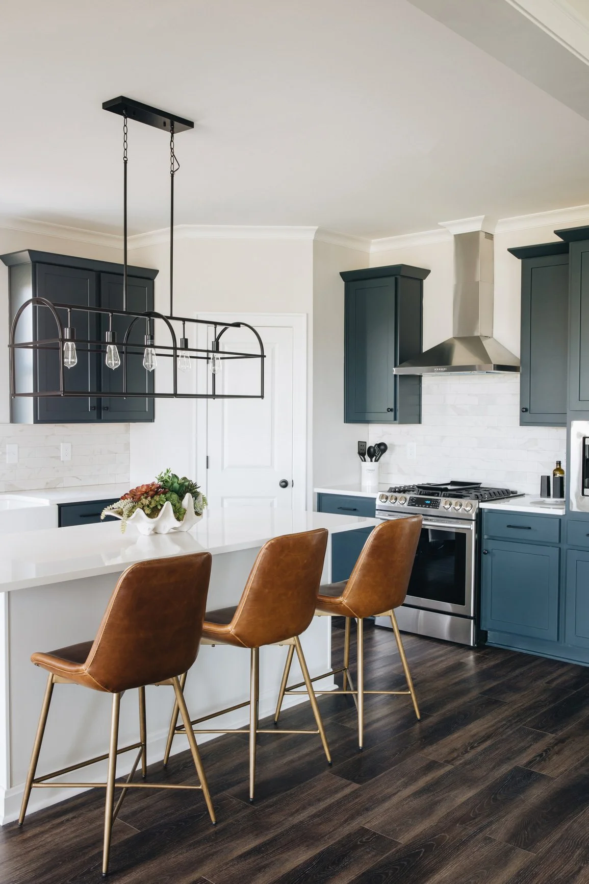 Modern kitchen with navy blue cabinets, white countertops, stainless steel oven and range hood, dark wood flooring, and three brown leather barstools at the island. There is a black lighting fixture above the island and a decorative white bowl with plants on the counter.
