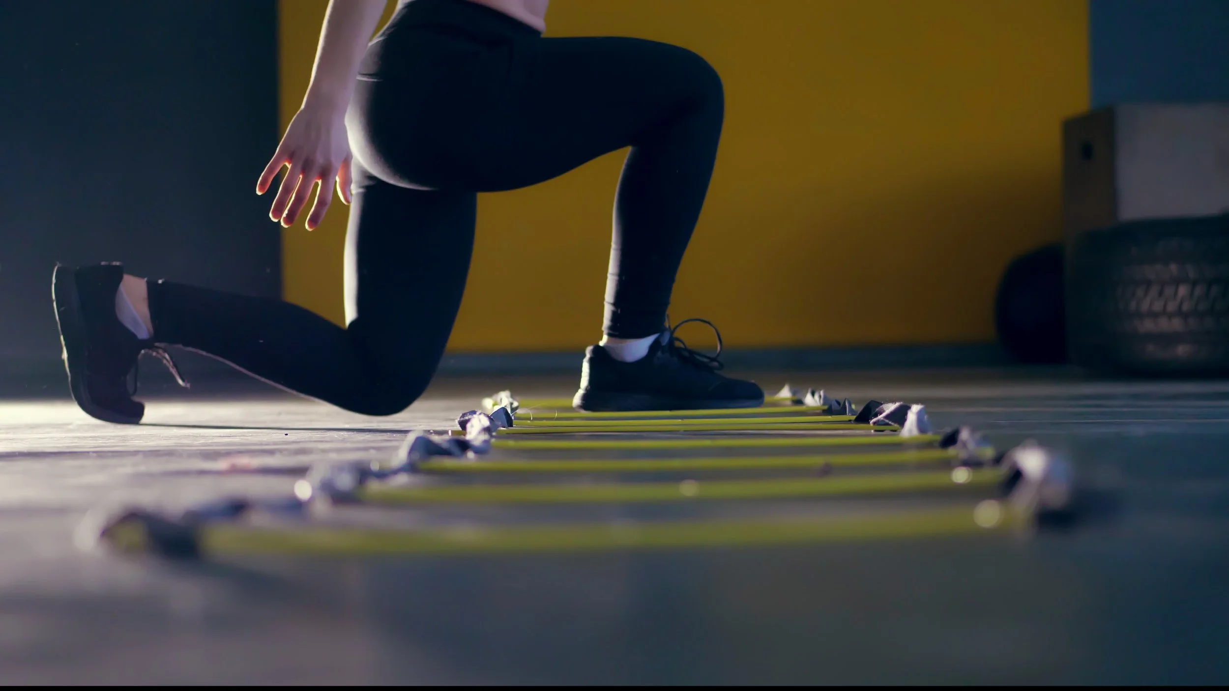 Person in athletic clothing performing a lunging exercise on a ladder on the gym floor.