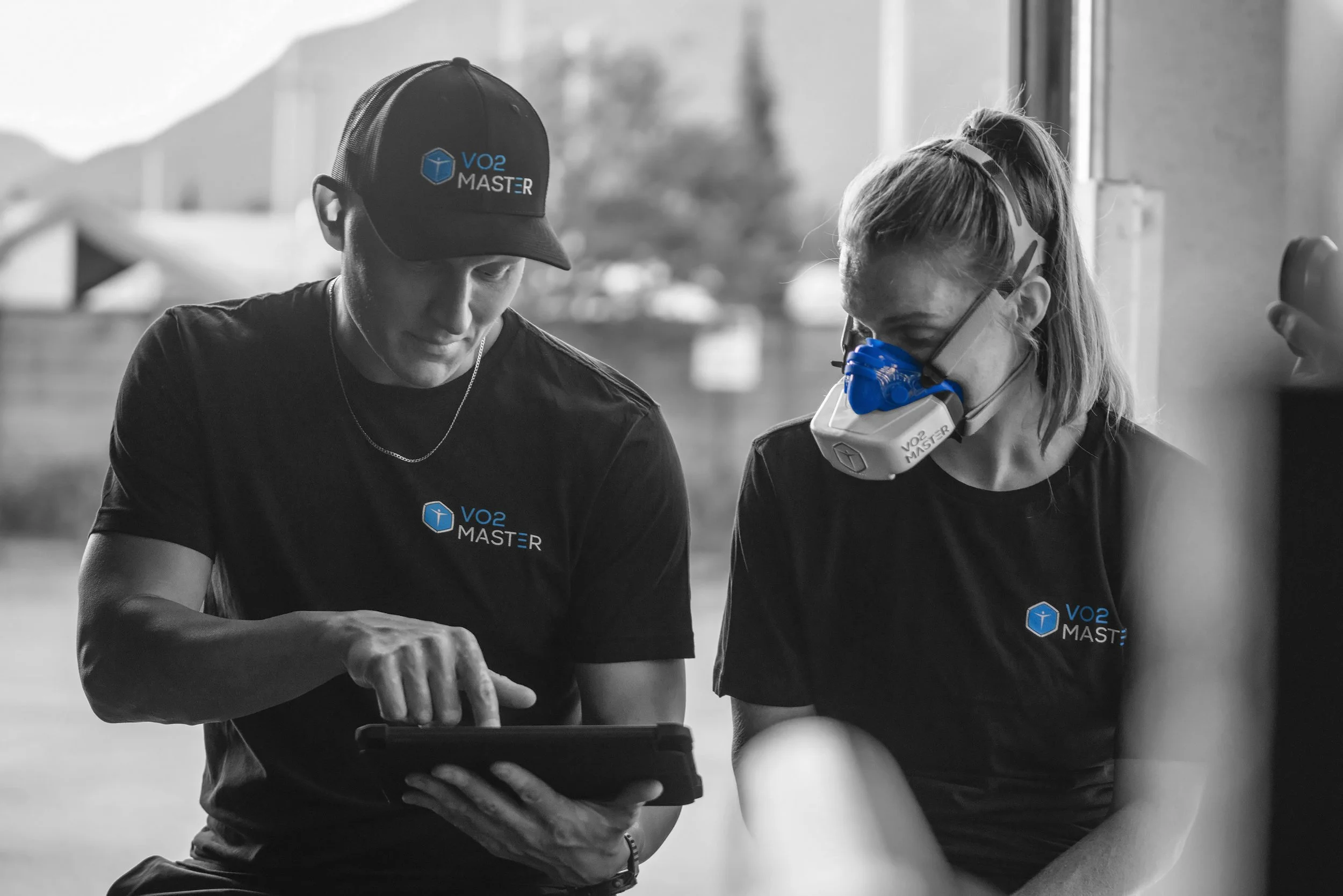 Two individuals wearing black shirts with a VO2 Master logo, one man with a cap and woman with a mask and protective eyewear, reviewing data on a tablet.