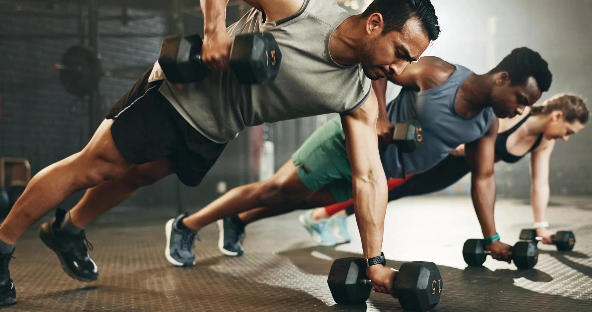 Three people doing push-ups with dumbbells in a gym.