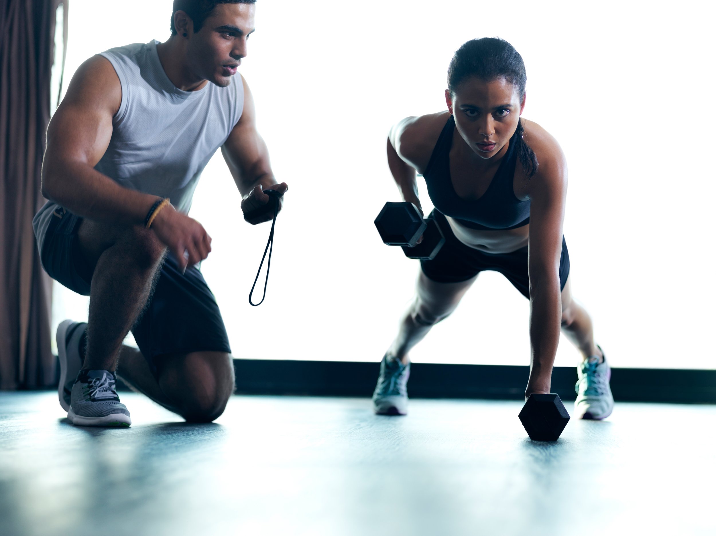 A woman doing a push-up with dumbbells under supervision of a trainer in a gym.