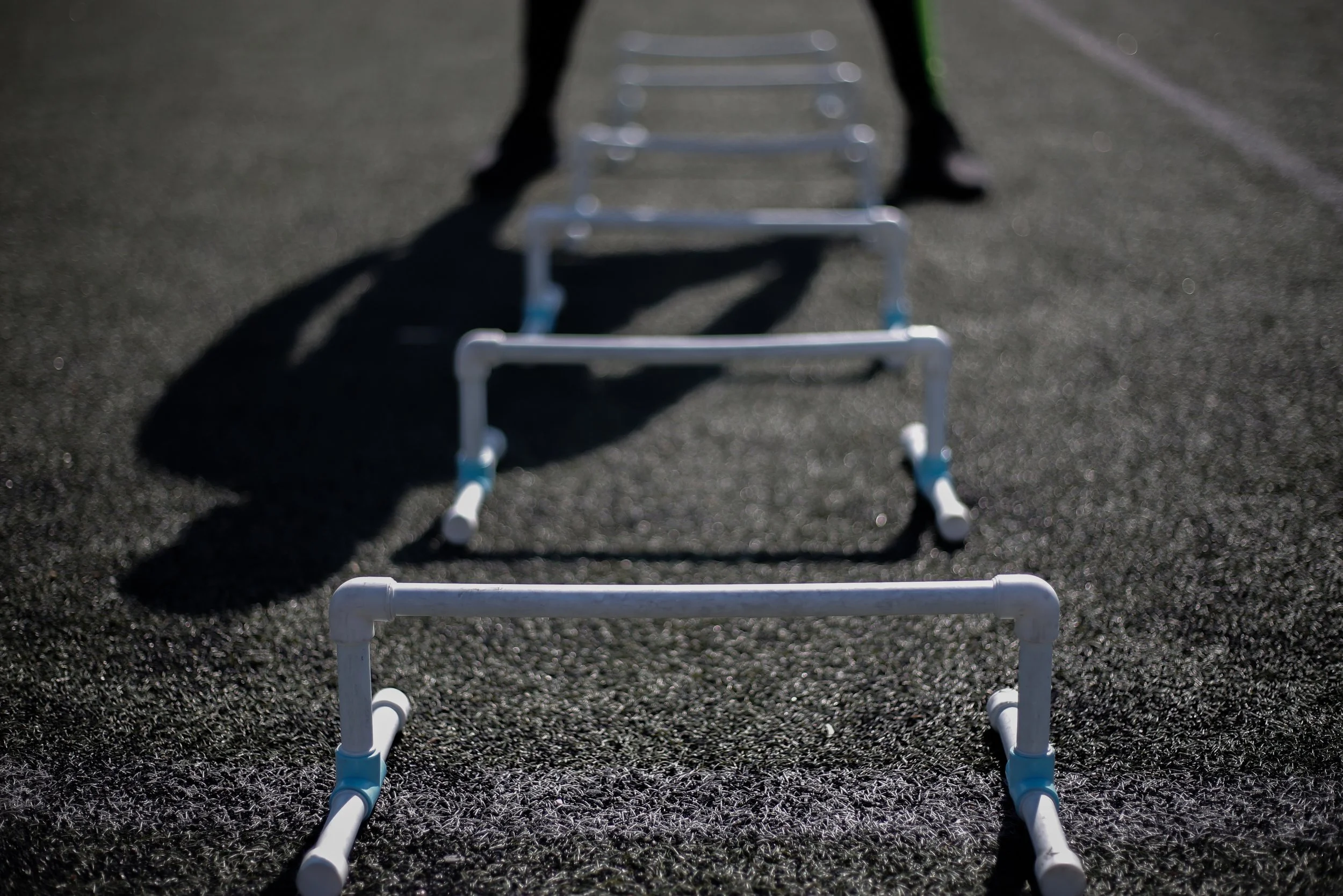 A row of white hurdle bars on a black outdoor running track with a person in dark clothing in the background.