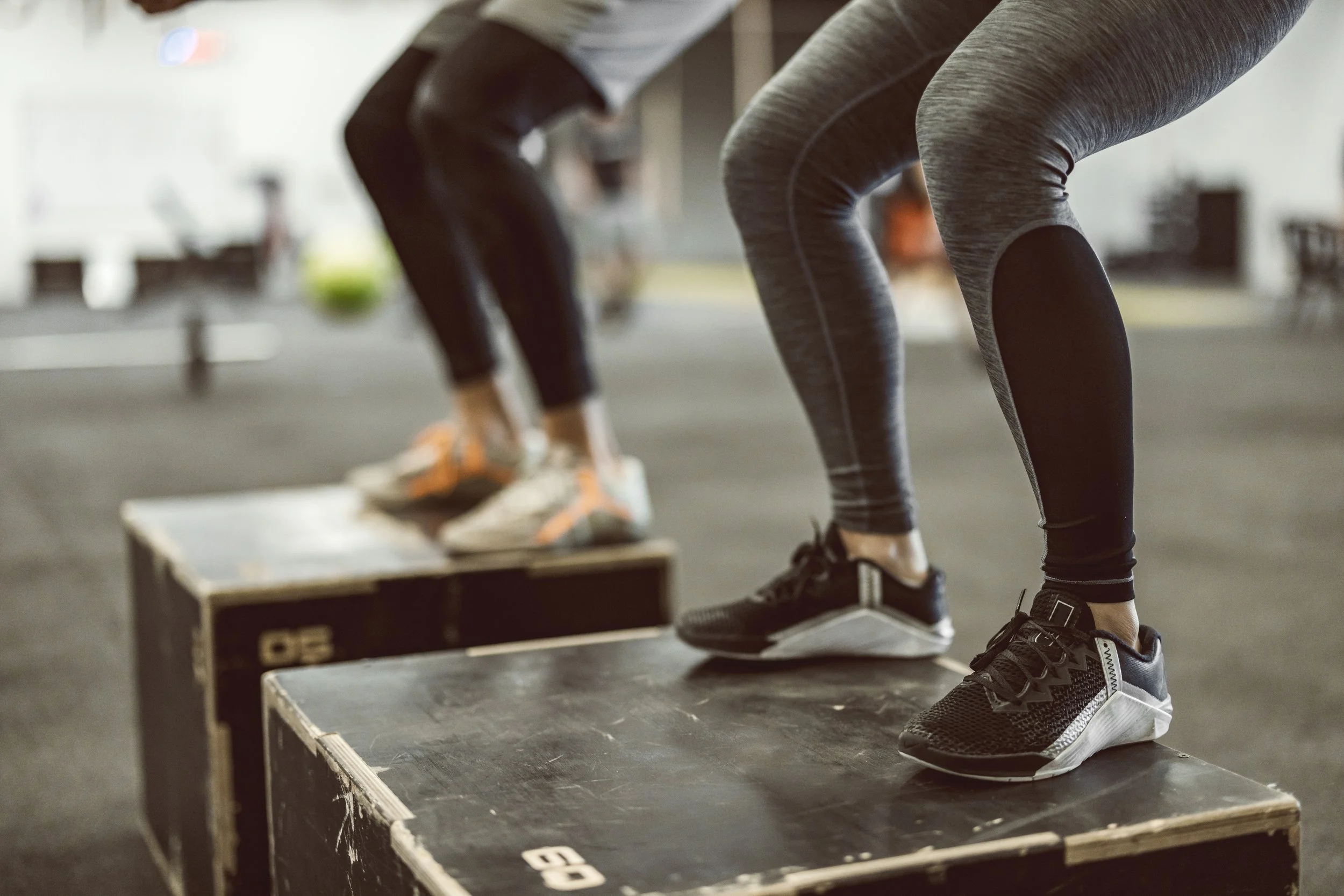 Two people exercising on wooden plyometric boxes in a gym, wearing athletic clothing and shoes, with one person in the foreground and the other in the background.