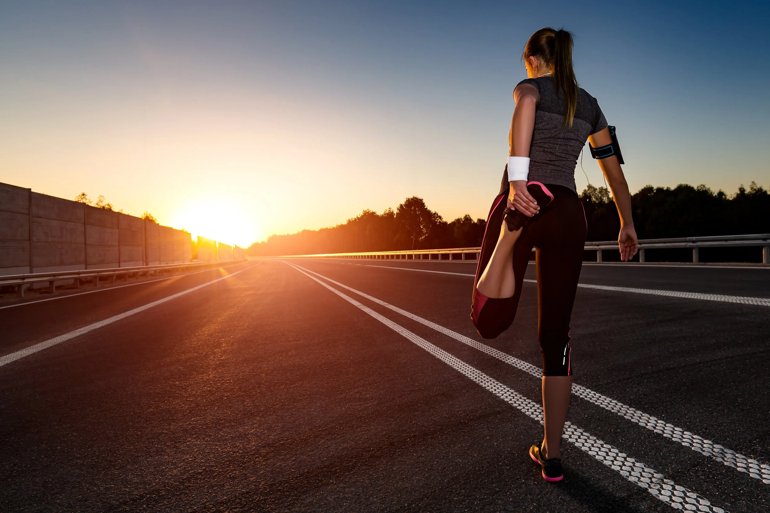 A woman stretching on the side of an empty road at sunrise or sunset, wearing workout clothes and an armband with a phone.