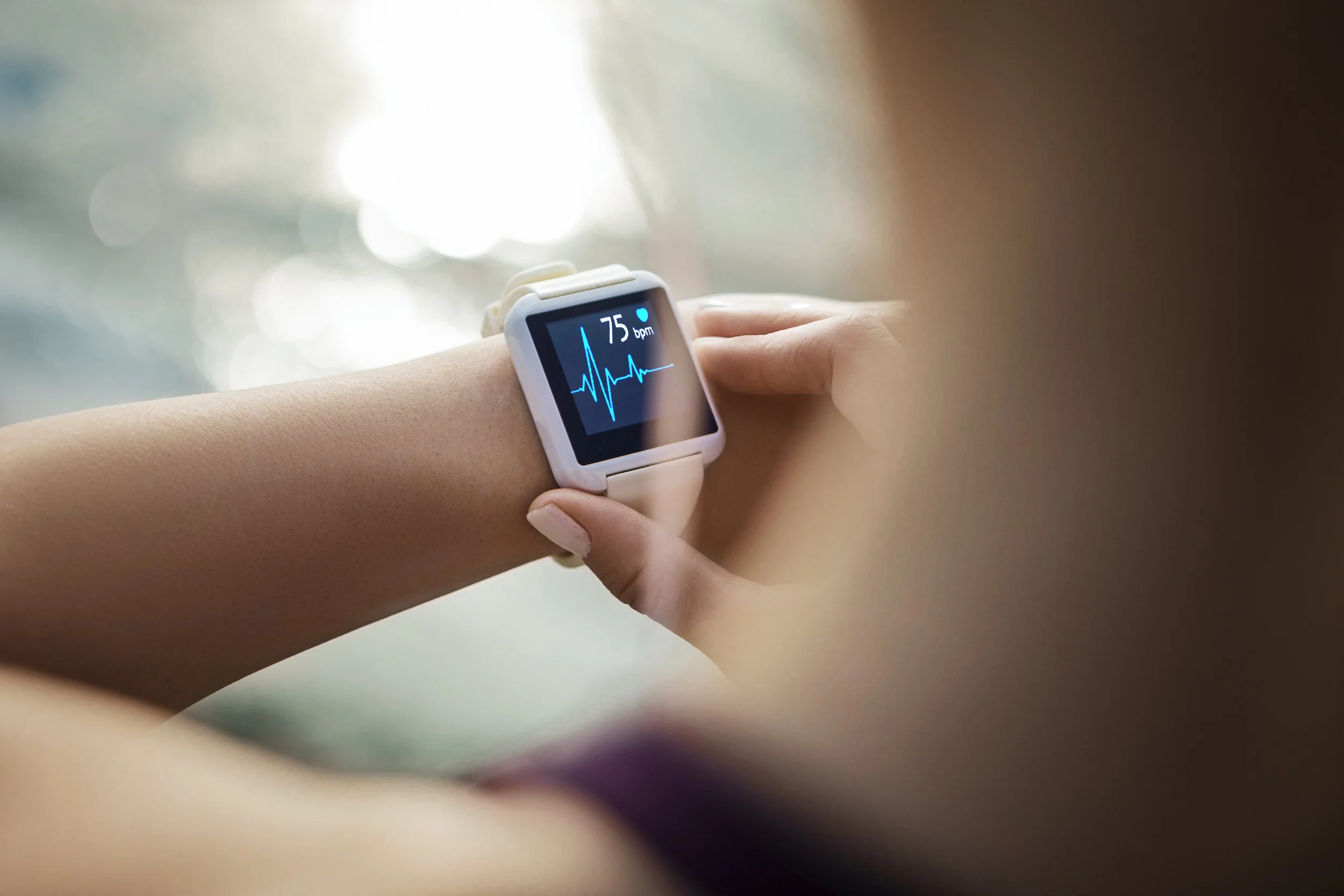 A person wearing a white smartwatch showing a heart rate of 75 bpm and an electrocardiogram on the display, with their hand touching their wrist.