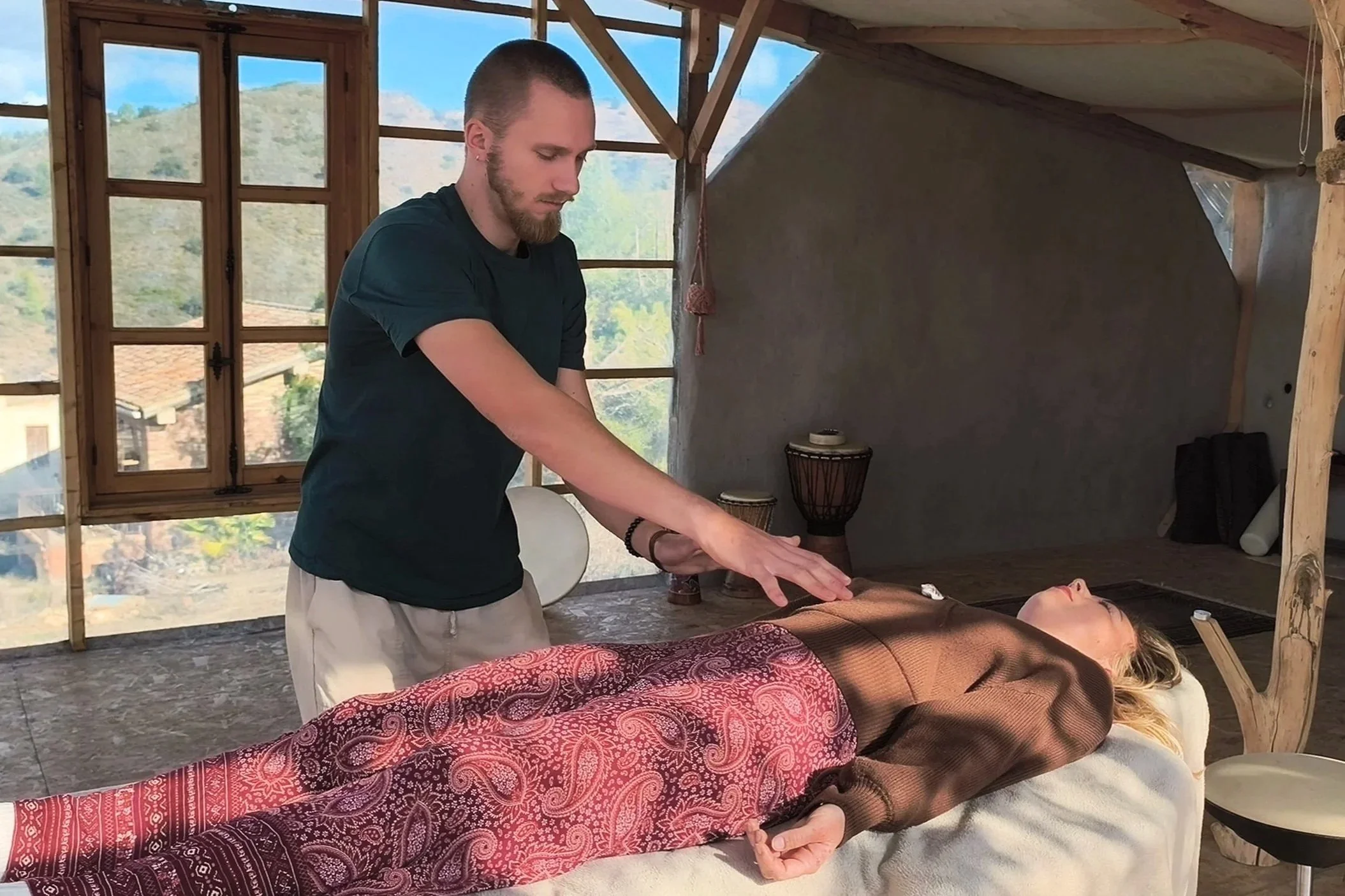 Ein Mann führt eine Reiki-Behandlung an einer liegenden Frau in einem Holzraum durch, mit großen Fenstern, die eine bergige Landschaft zeigen.