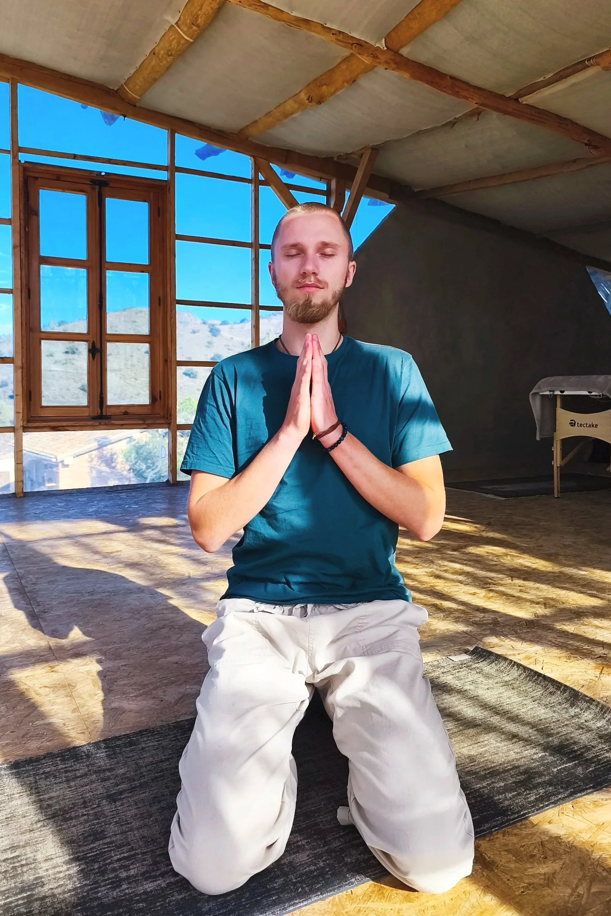 Ein junger Mann mit geschlossenen Augen in einer Meditationshaltung im Innenraum eines Hauses mit großen Fenstern, Holzdecke und Holzboden, bei Sonnenlicht.
