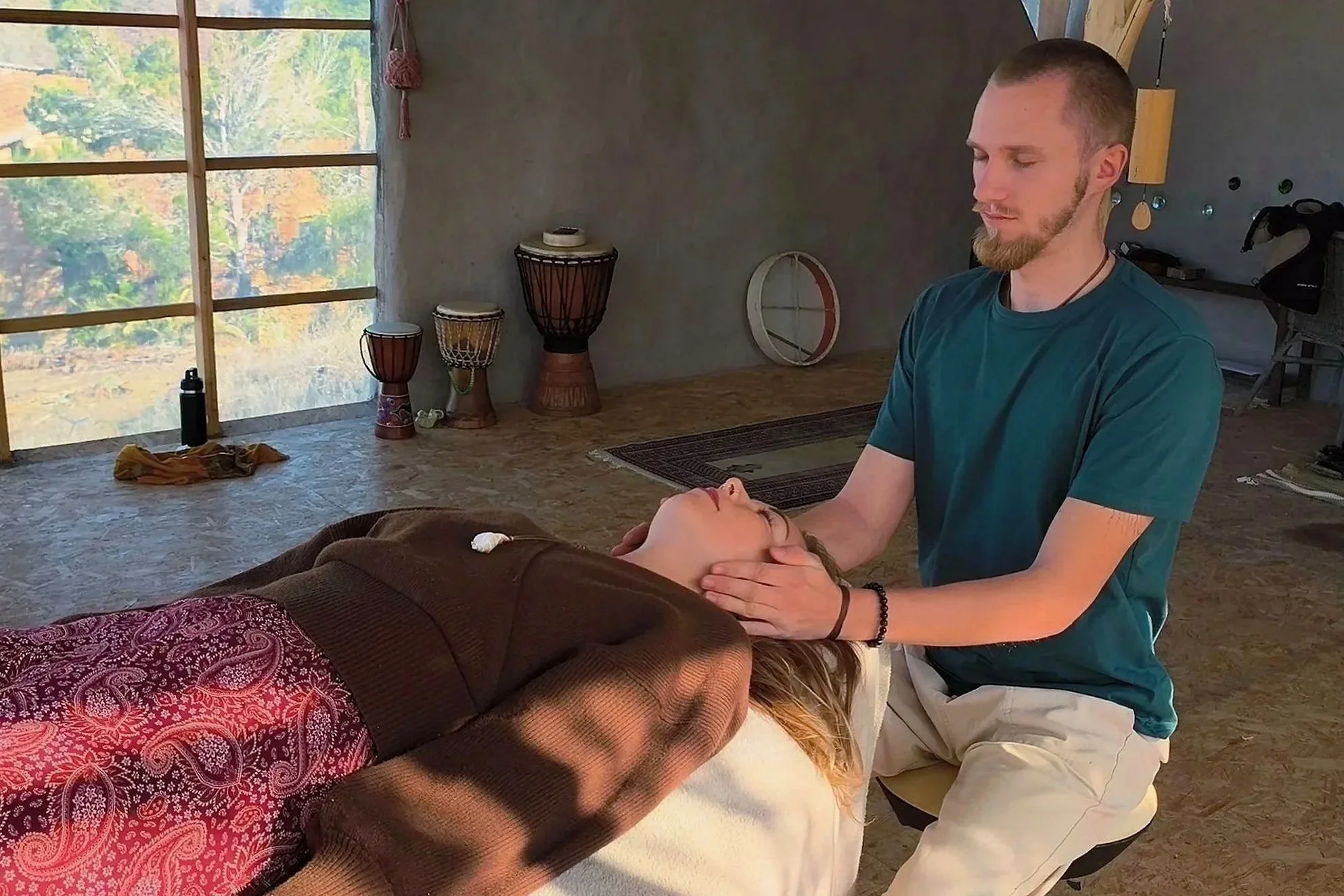 Ein Mann führt eine Therapie oder Heilbehandlung bei einer liegenden Frau durch, die entspannt das Gesicht hält, in einem Raum mit großen Fenstern und Dattelpalmenschalen an der Wand.