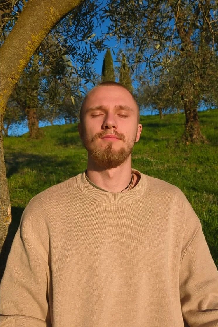 Junger Mann mit geschlossener Augen entspannend im Sonnenschein, im Freien unter Bäumen mit grünem Rasen im Hintergrund.