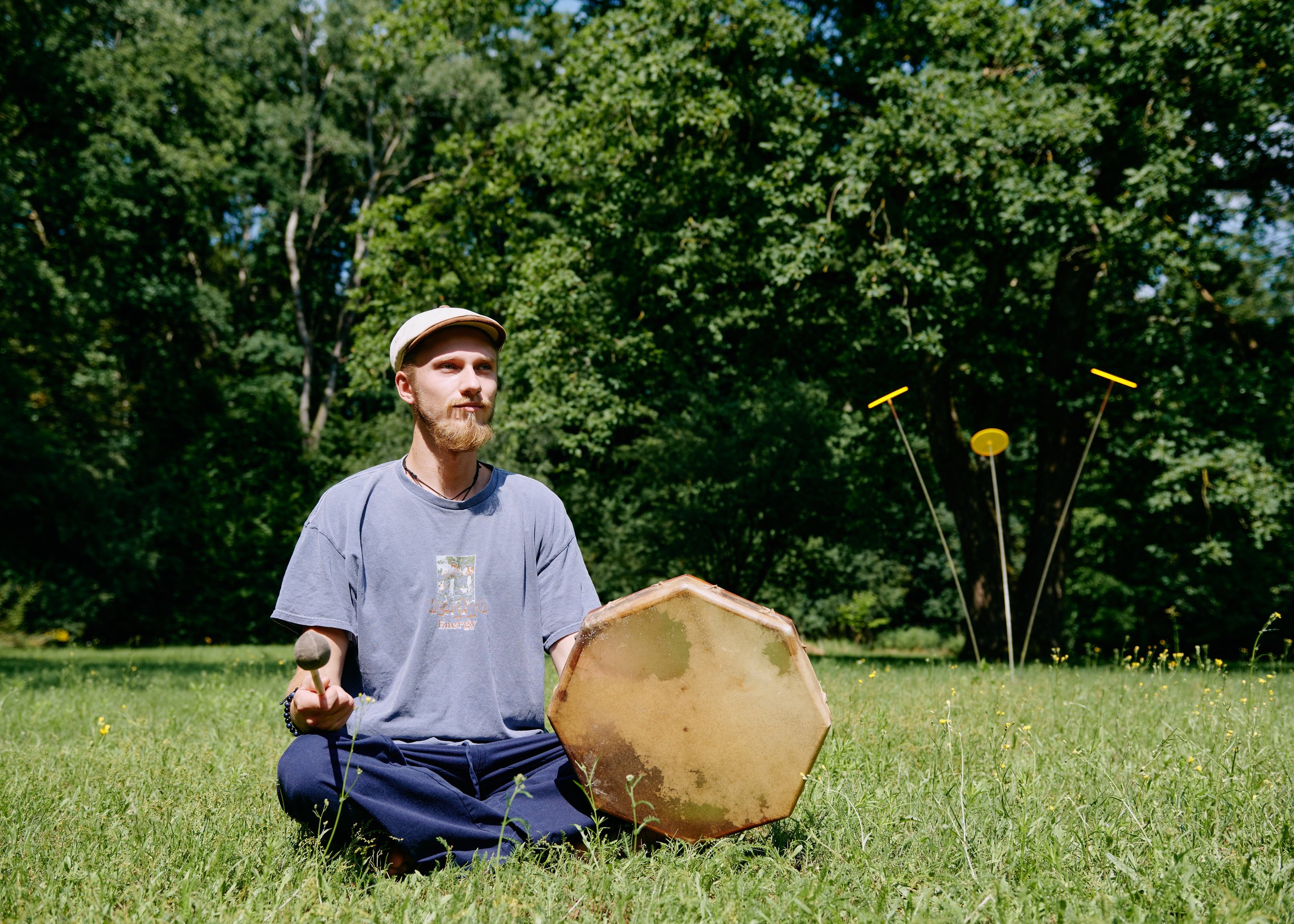 Ein Mann sitzt kreuzbein auf einer Wiese, spielt eine Trommel und schaut nach vorne, im Hintergrund befinden sich Bäume und zwei Deko-Objekte in Form von Teleskopstangen mit Scheiben und Stäben.