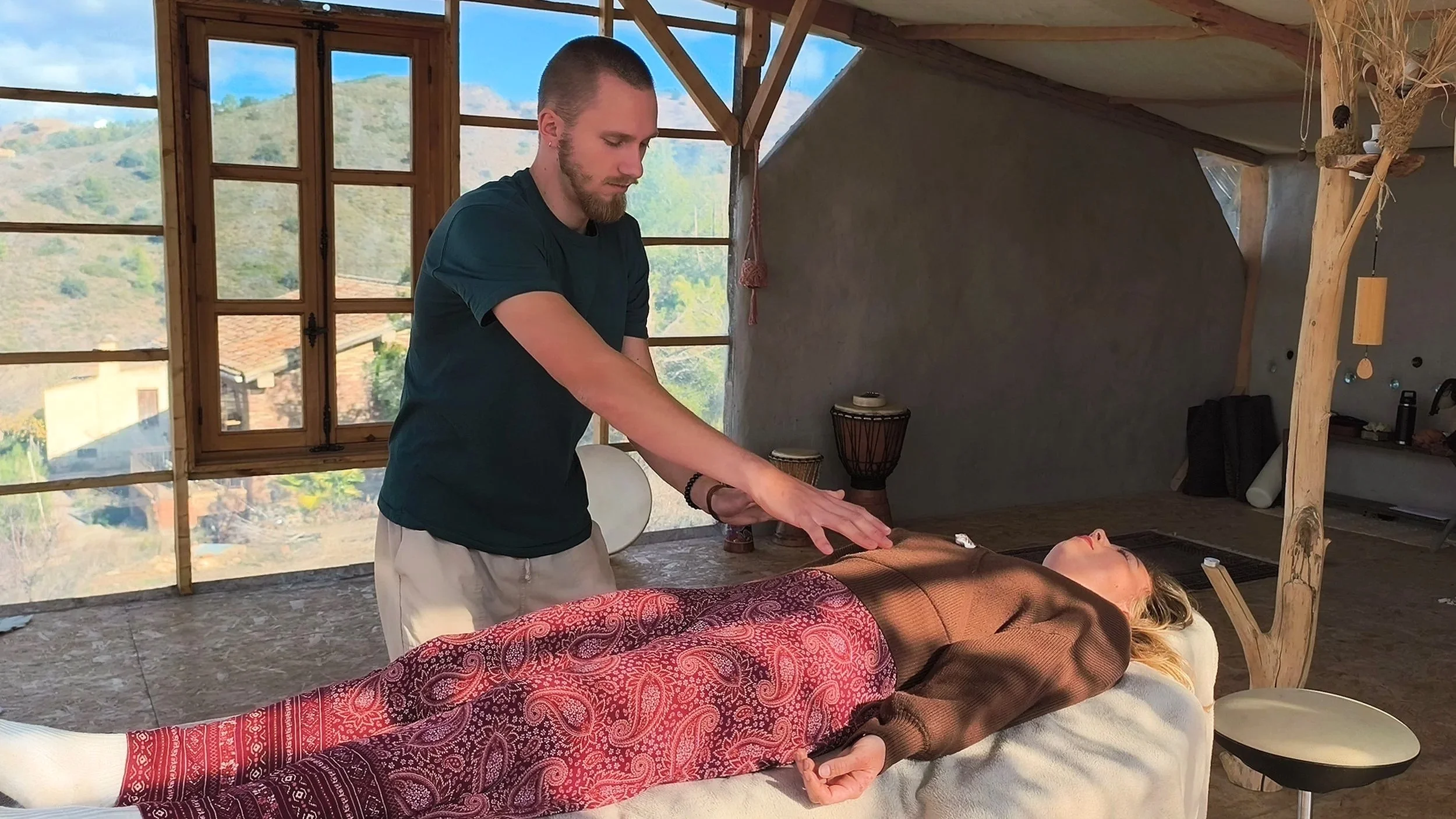 Eine Person liegt auf einer Massagebank, während eine andere Person eine Energiebalance oder Heilbehandlung durchführt, in einem Raum mit großen Fenstern und Holzelementen.