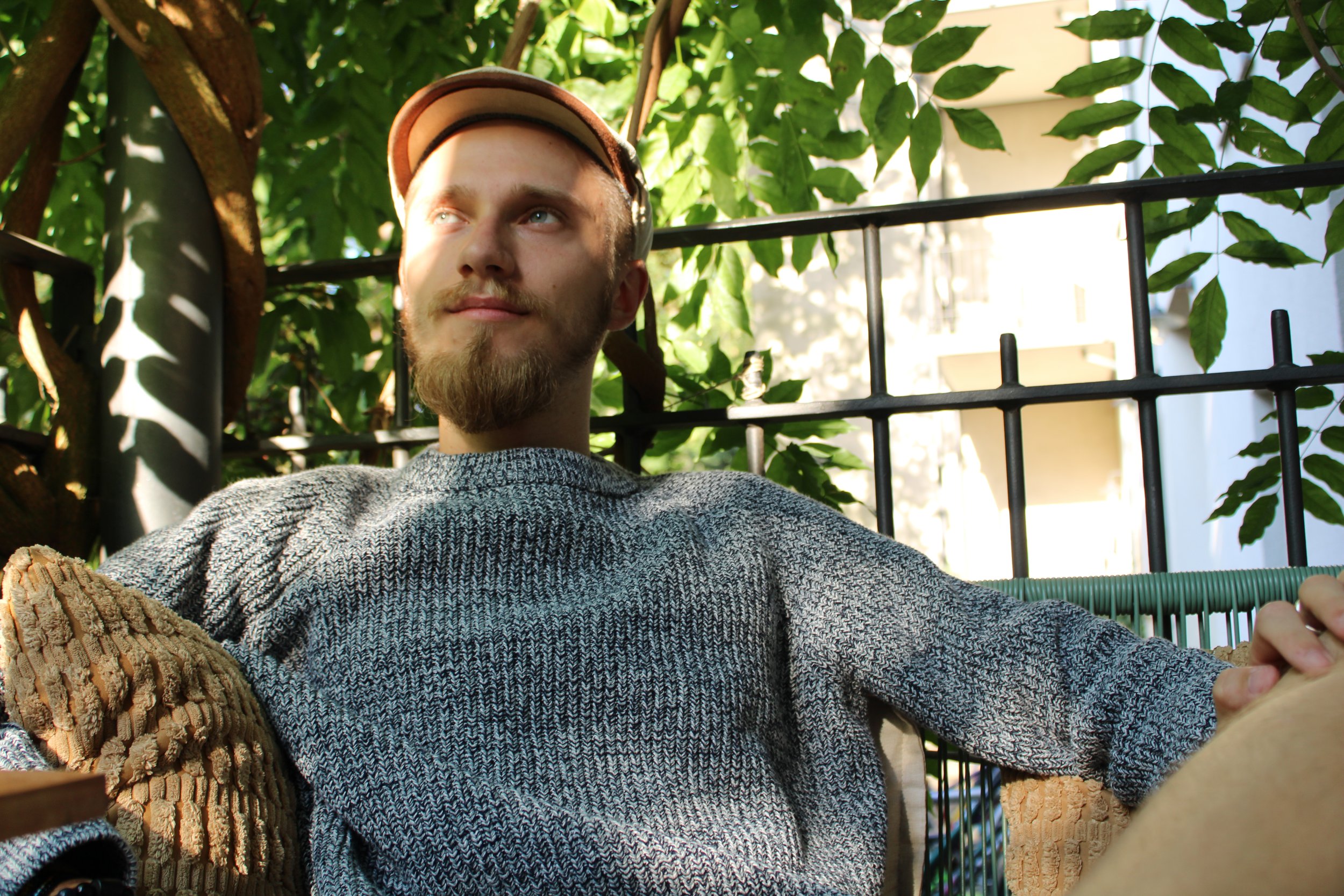 Ein junger Mann mit Bart sitzt entspannt auf einem Balkon, umgeben von grünen Pflanzen und Sonnenschein.