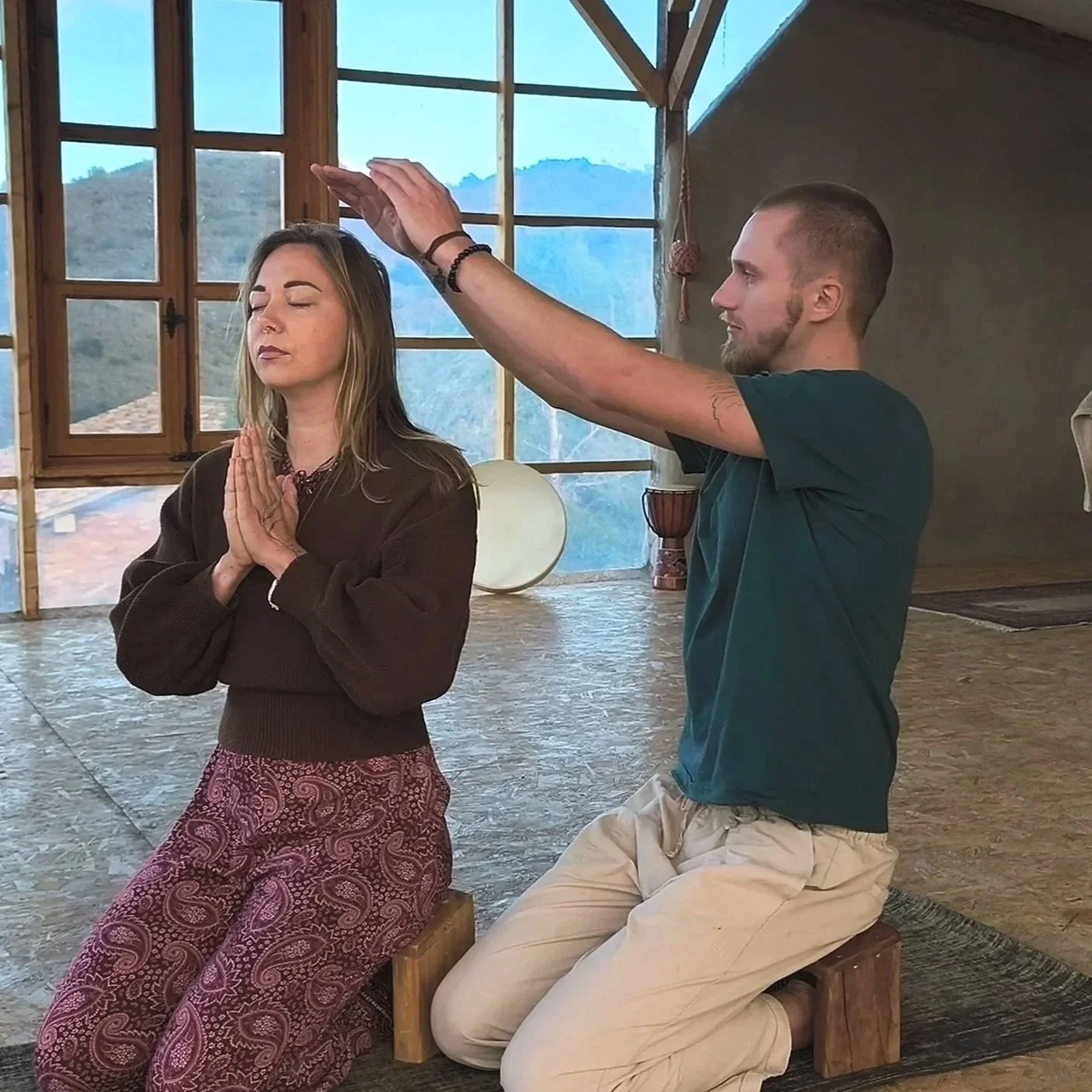 Zwei Menschen sitzen im Meditationsraum, der Mann führt eine Heilungsübung durch, die Frau sitzt mit geschlossenen Augen und in meditativer Haltung.