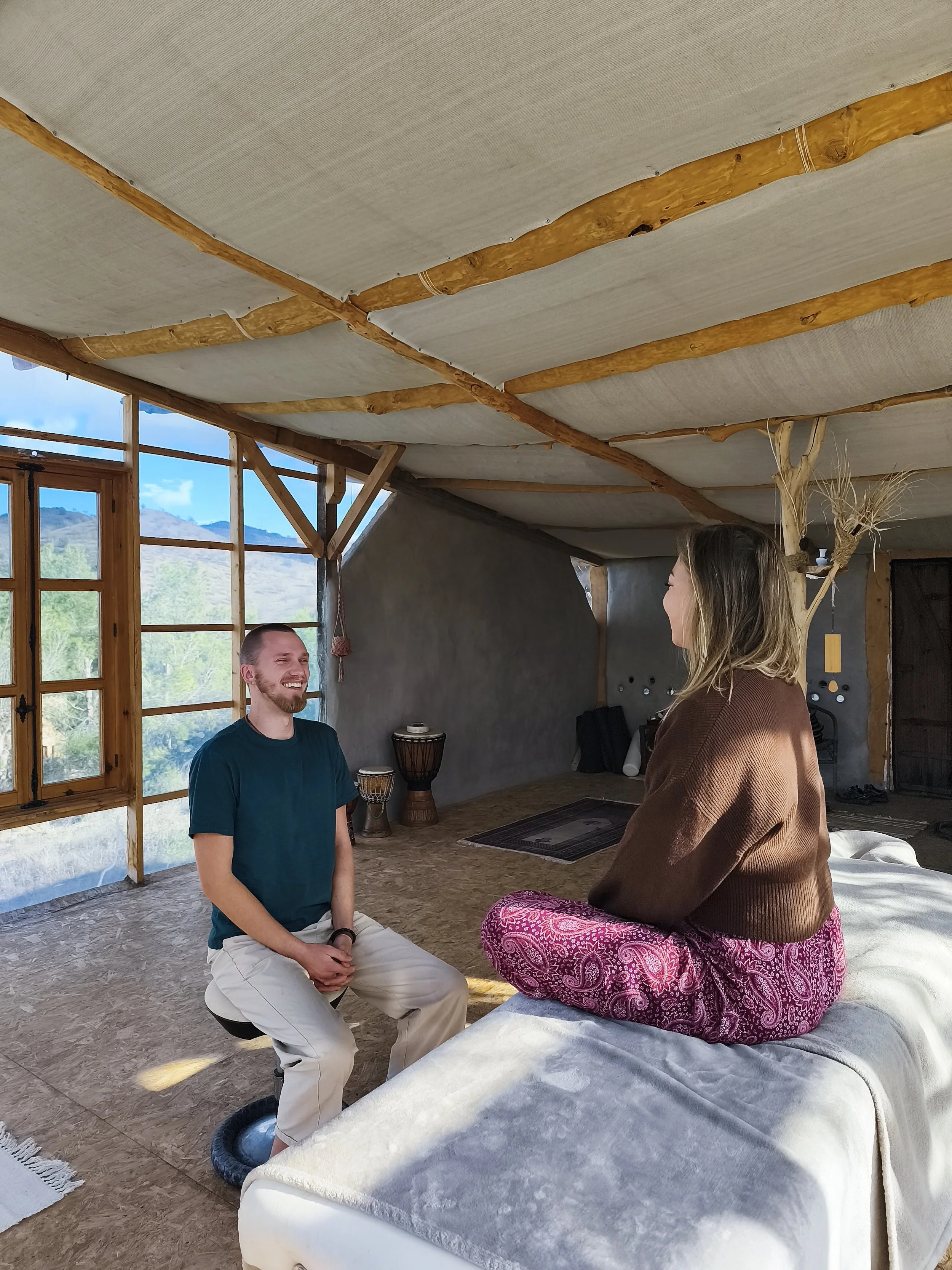 Zwei Menschen sitzen im Raum, lachen und sprechen miteinander, mit einem warmen, natürlichen Stil, Holzdecke, große Fenster und Vitaminelemente im Hintergrund.